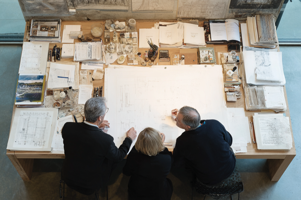 post thumbnails - Deux hommes et une femme, tous habillés en noir, regarde des plans d'architecture déposés sur une grande table en bois. Des dessins et plans sont affichés sur le mur en béton.