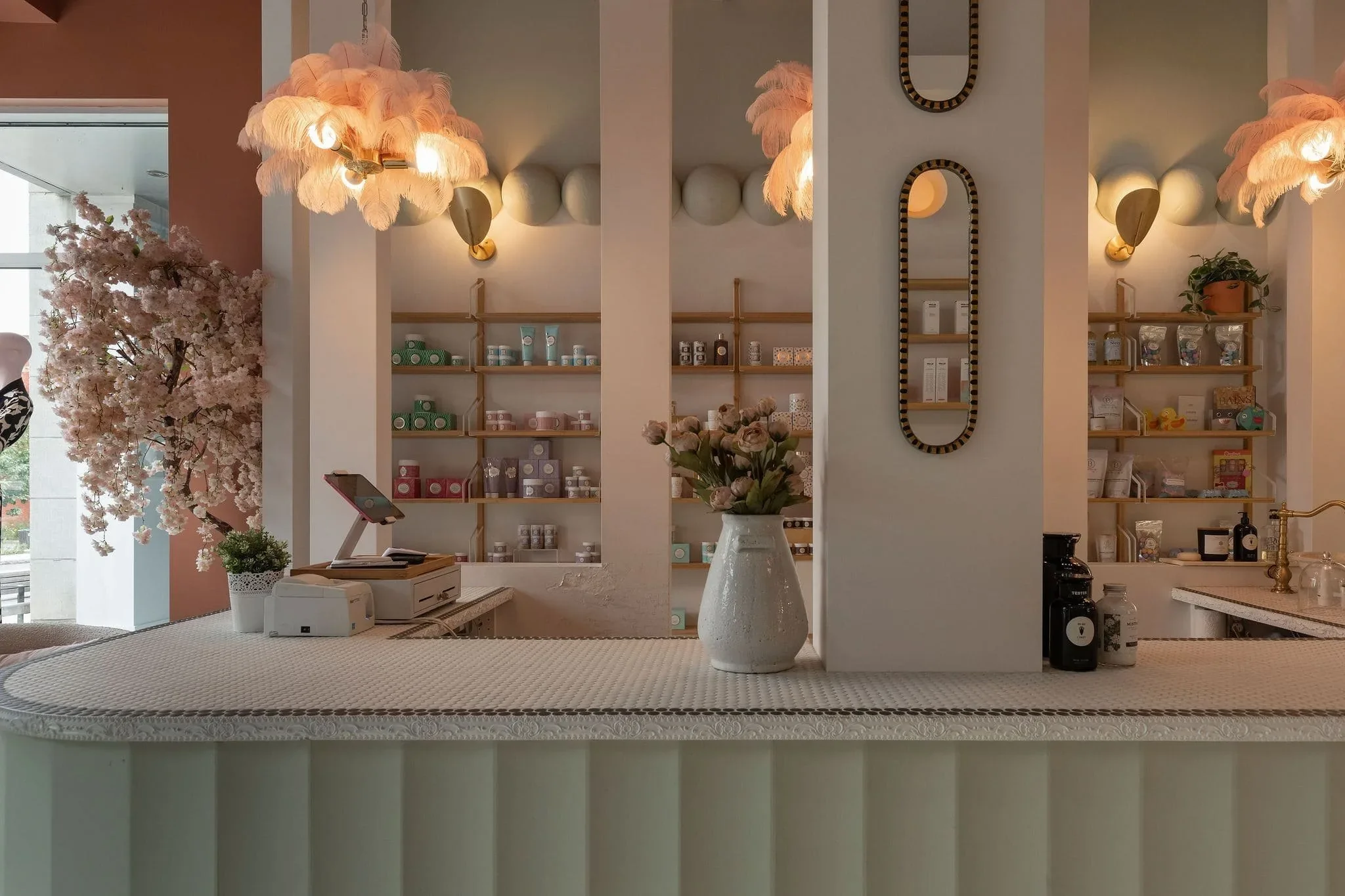 Comptoir de boutique à Montréal par Rübik Design : luminaires en plumes roses, colonnes blanches, miroirs allongés et étagères en bois