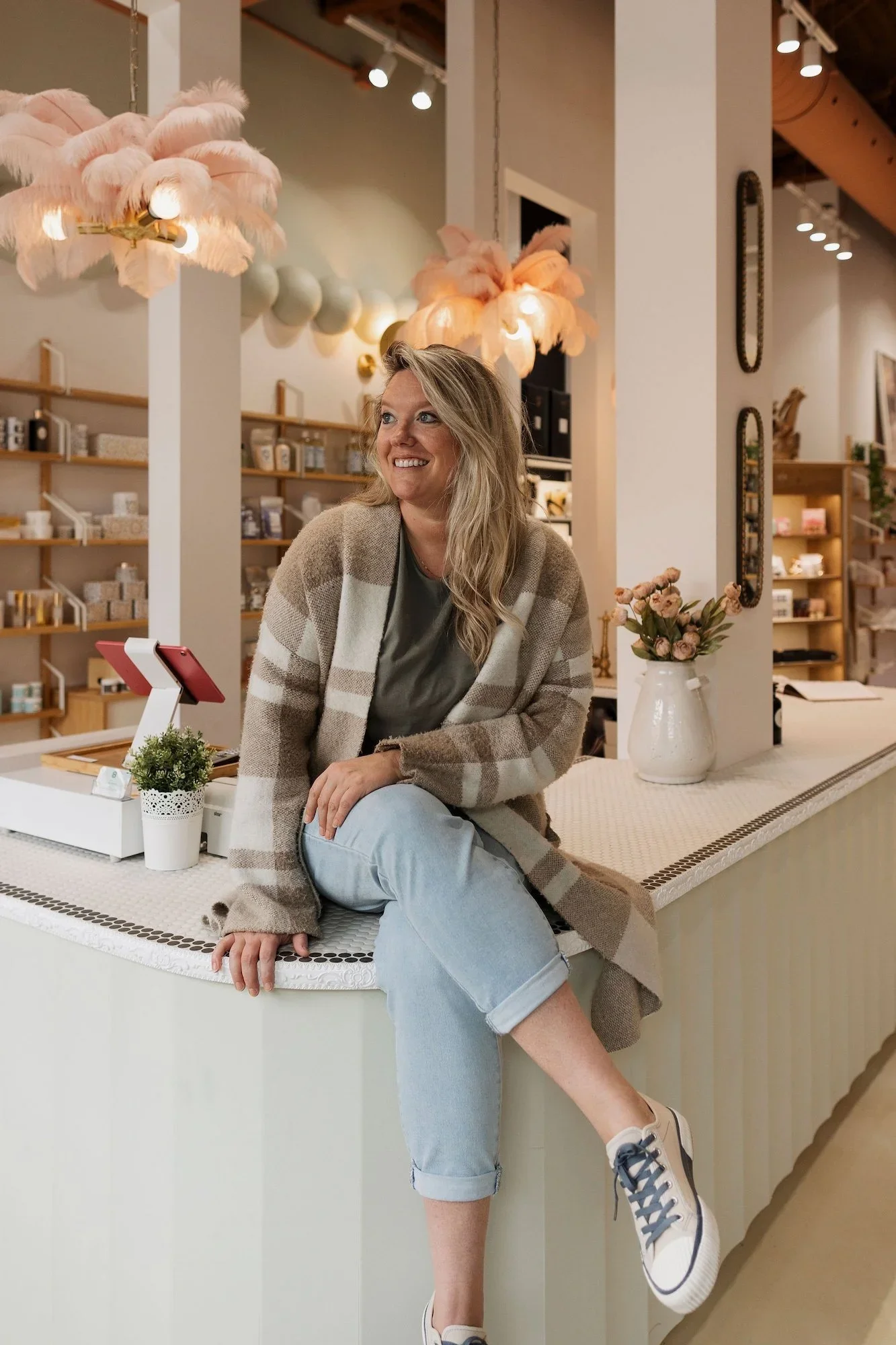 Sophie Roy, fondatrice et directrice créative de RÜBIK DESIGN, assise sur un comptoir de boutique sous des luminaires à plumes roses