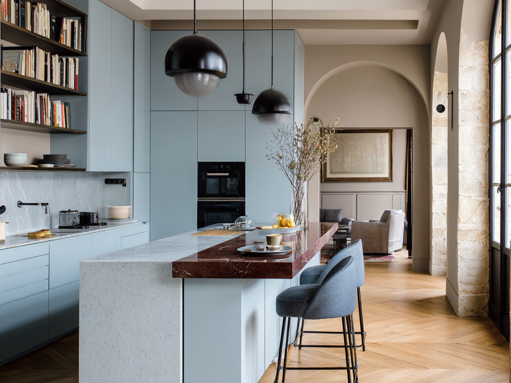 Cuisine moderne avec des armoires de rangement bleu clair, et des étagères ouvertes avec libres et vaisselles, un ilôt central en marbre avec un accent marron, deux tabourets de bar bleu marine et des lampadaires suspendus noirs.