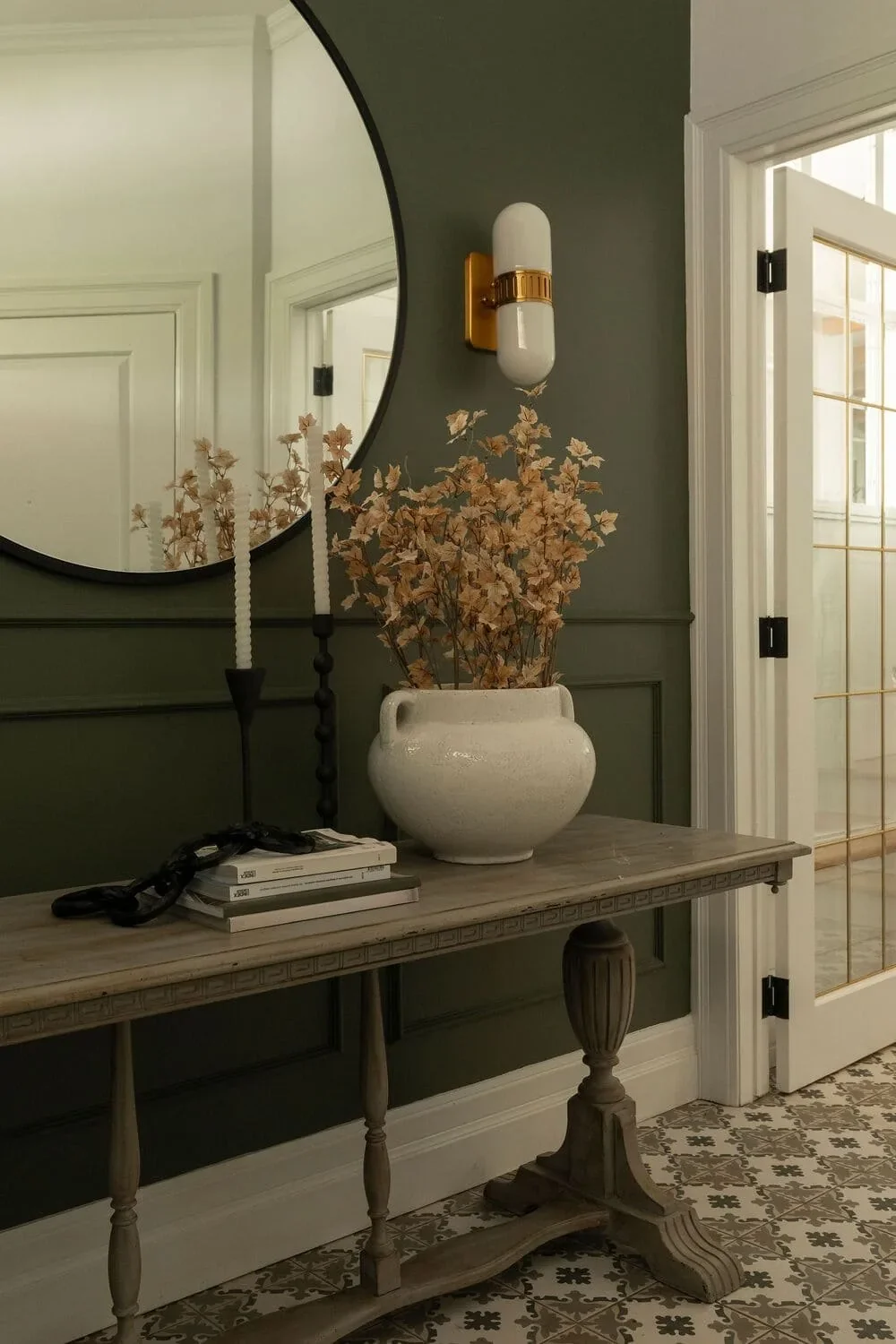 Entrée d'une maison de la Rive-Nord par Rübik Design : mur vert olive, miroir rond, console en bois, applique dorée et vase de fleurs séchées