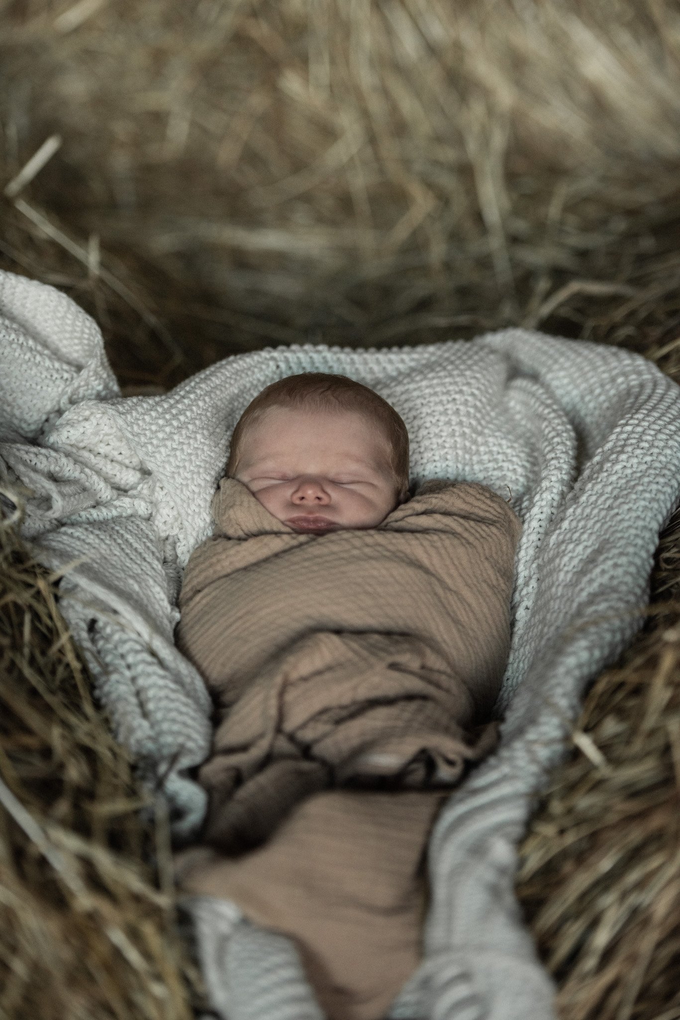 Close-up of a newborn baby sleeping in a crib made of straw, wrapped in a tan blanket, with a white knitted blanket surrounding them.