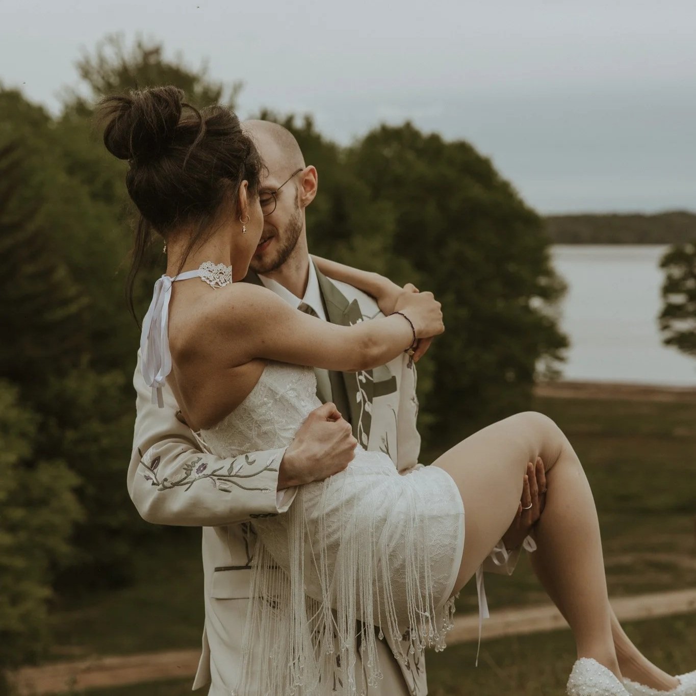 A man is lifting a woman who is wearing a wedding dress outdoors near a body of water and trees, sharing an intimate moment.