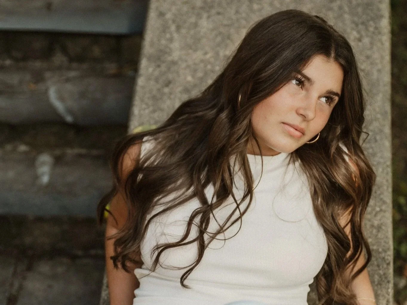 Young woman with long wavy dark hair, wearing a white top and gold hoop earrings is sitting outdoors next to a stone wall.