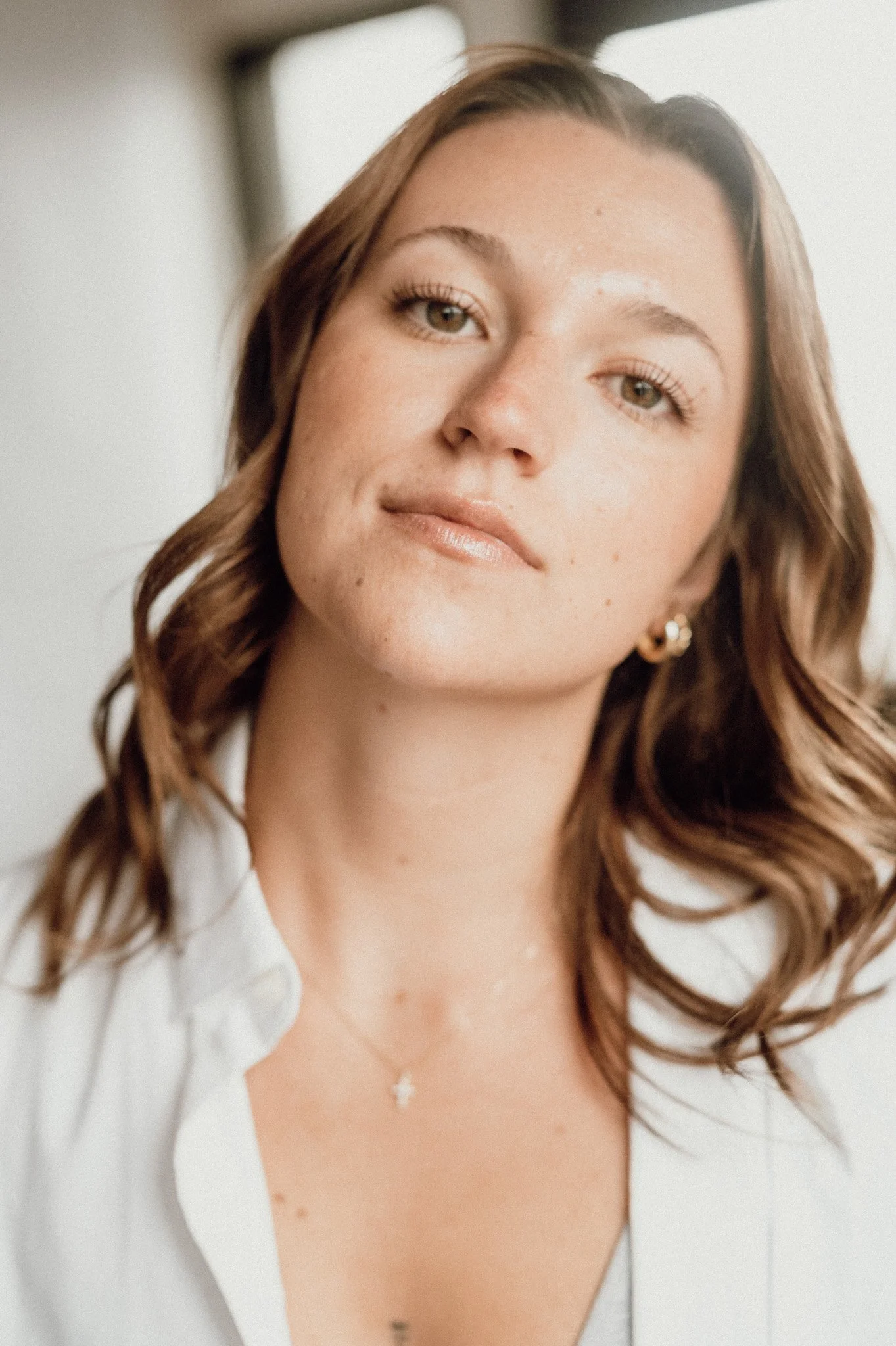 A young woman with light brown hair styled in loose waves, wearing a white top with a V-neck and small earrings, standing against a plain light background.