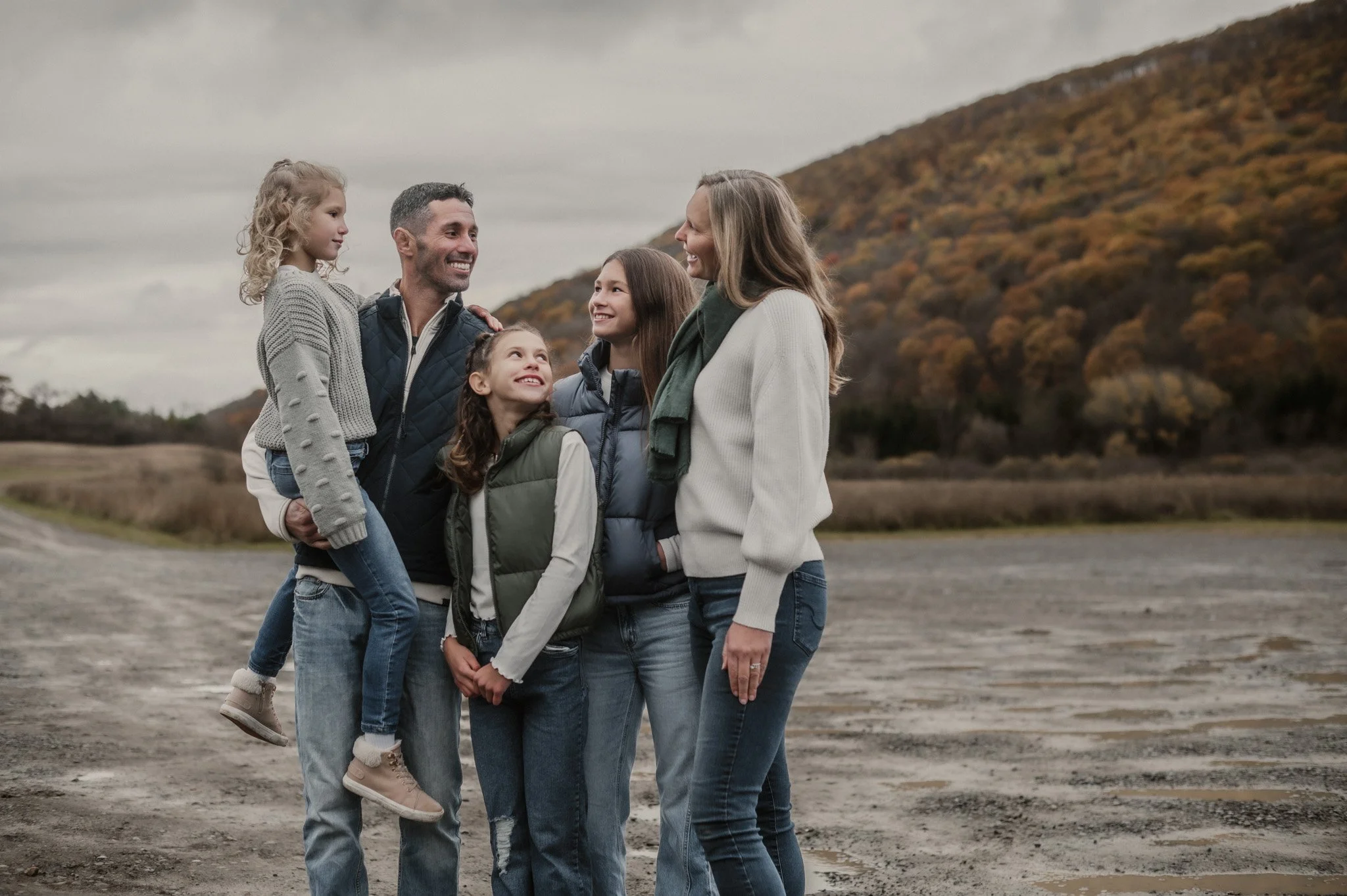 A family of five stands outdoors on a dirt path with autumn trees and hills in the background, smiling and looking at each other.