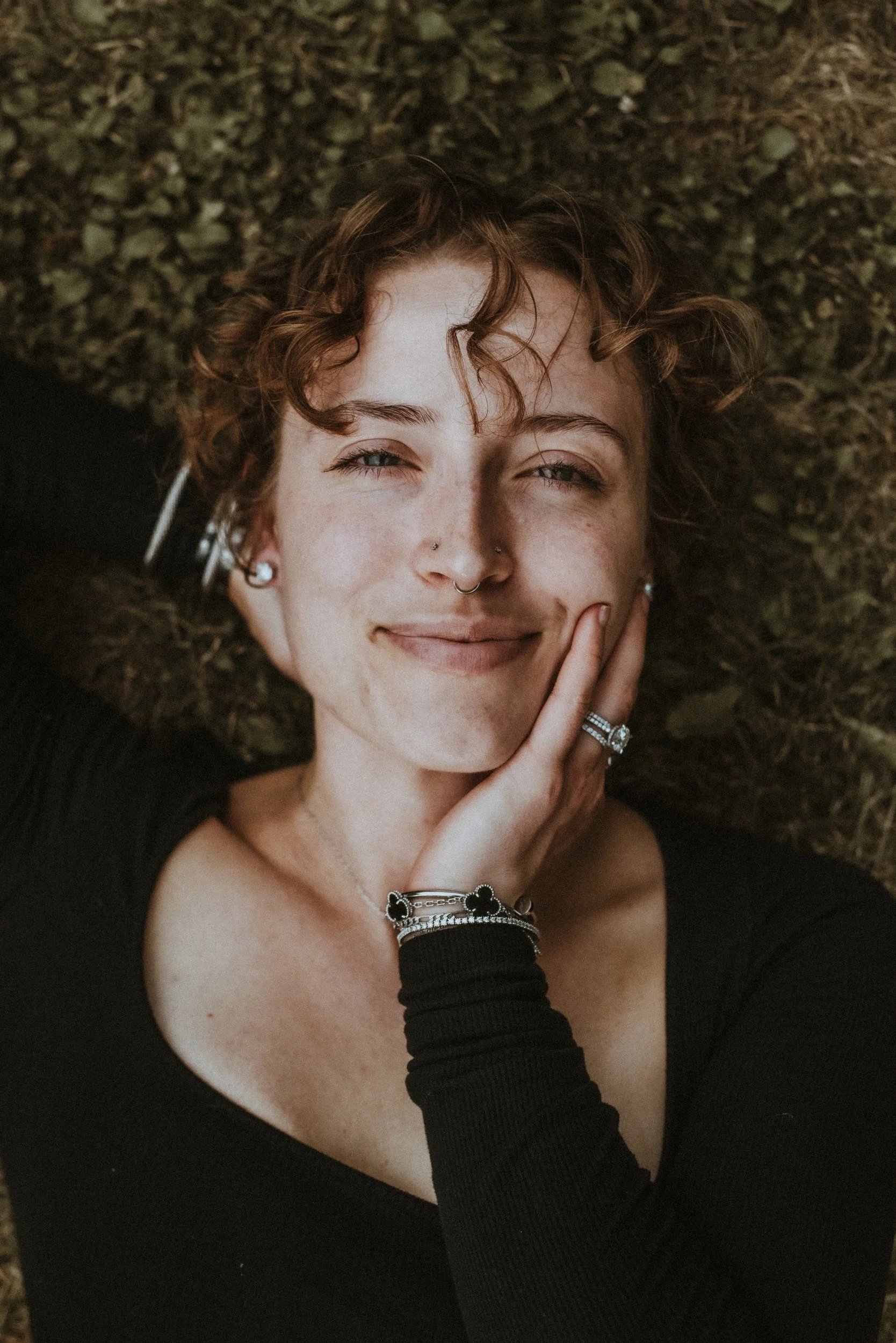 A woman with curly short hair, piercings, and jewelry lying on a textured dark surface, smiling and touching her face.