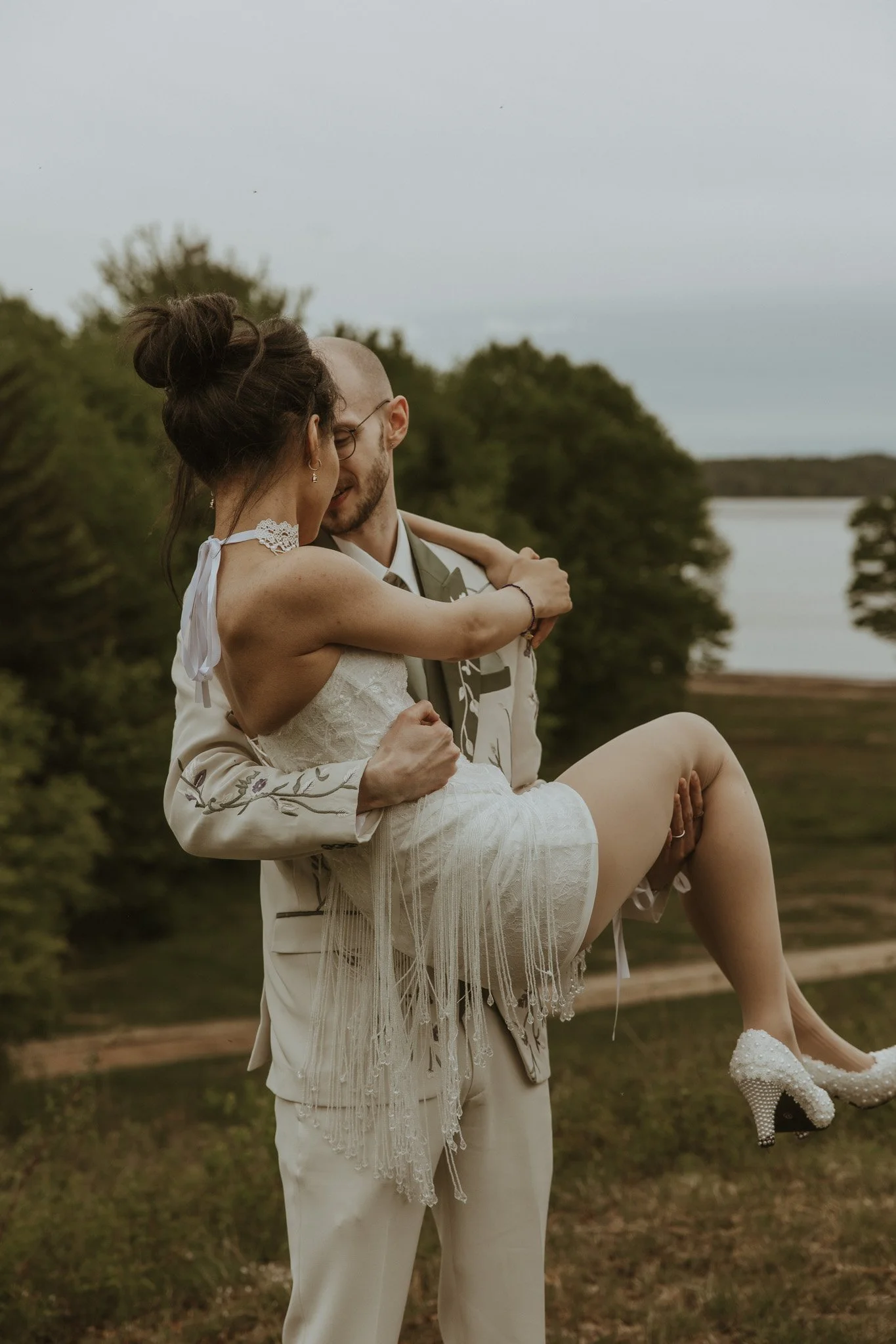 A man dressed in a beige suit holds a woman in a white dress and pearl high heel shoes during a wedding celebration outdoors by a river with trees in the background.