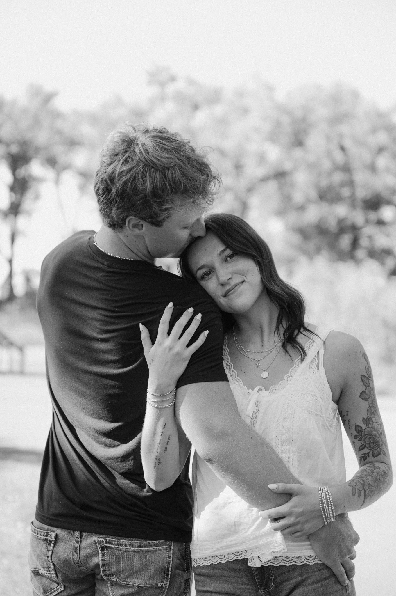 A black and white photo of a couple embracing outdoors. The man is kissing the woman on the forehead, and the woman is resting her head on his chest, smiling softly. The woman has tattoos on her arms and wearing a white lace dress.