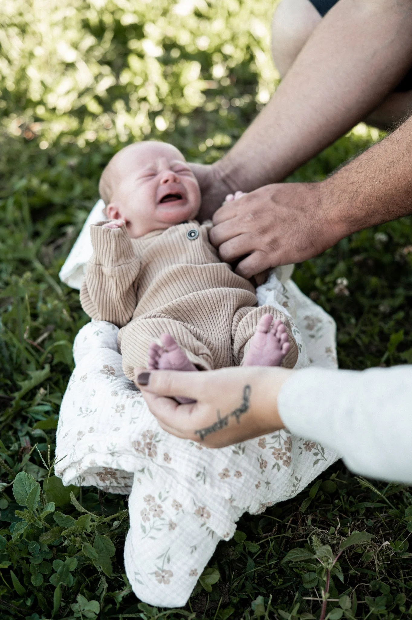 A crying baby lying on a white blanket on the grass, being comforted by an adult with visible tattooed hands, outdoors with greenery in the background.