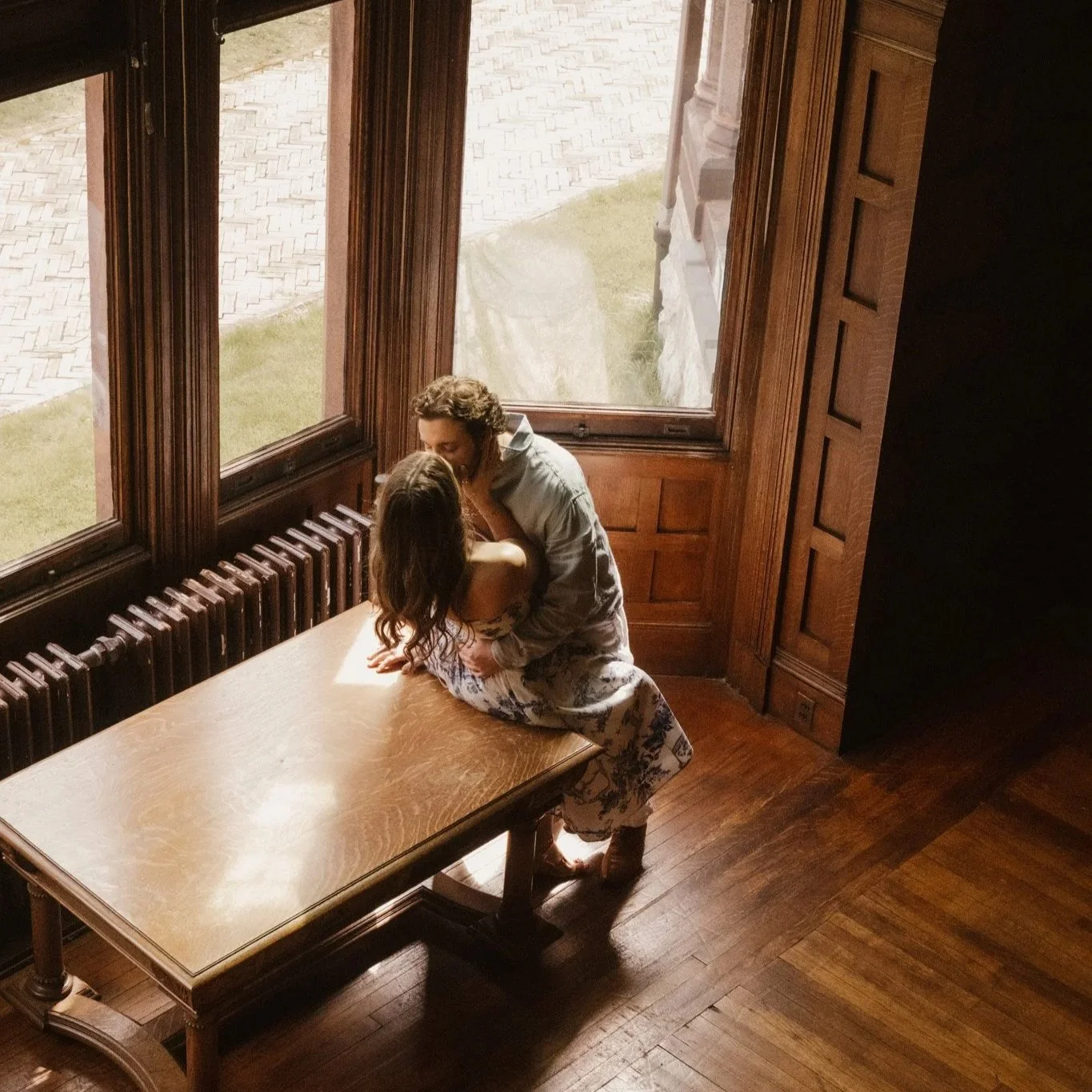A couple sitting at a wooden table near large windows, embracing and sharing a kiss in a cozy room with wooden floors and paneling.