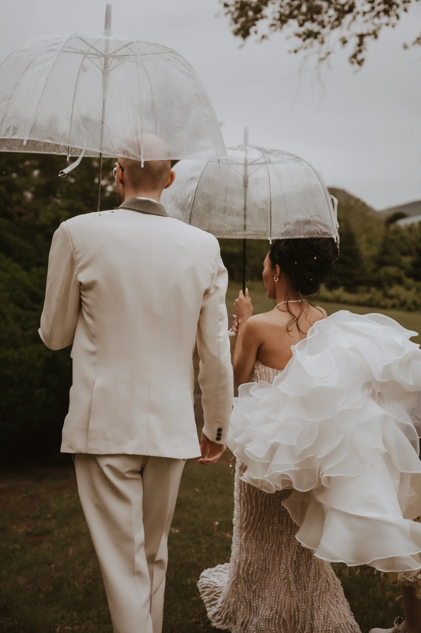 A newly married couple holding hands and walking outdoors on a rainy day, each holding a transparent umbrella. The bride is wearing a wedding dress with ruffles, and the groom is in a white suit. The background shows trees and a cloudy sky.