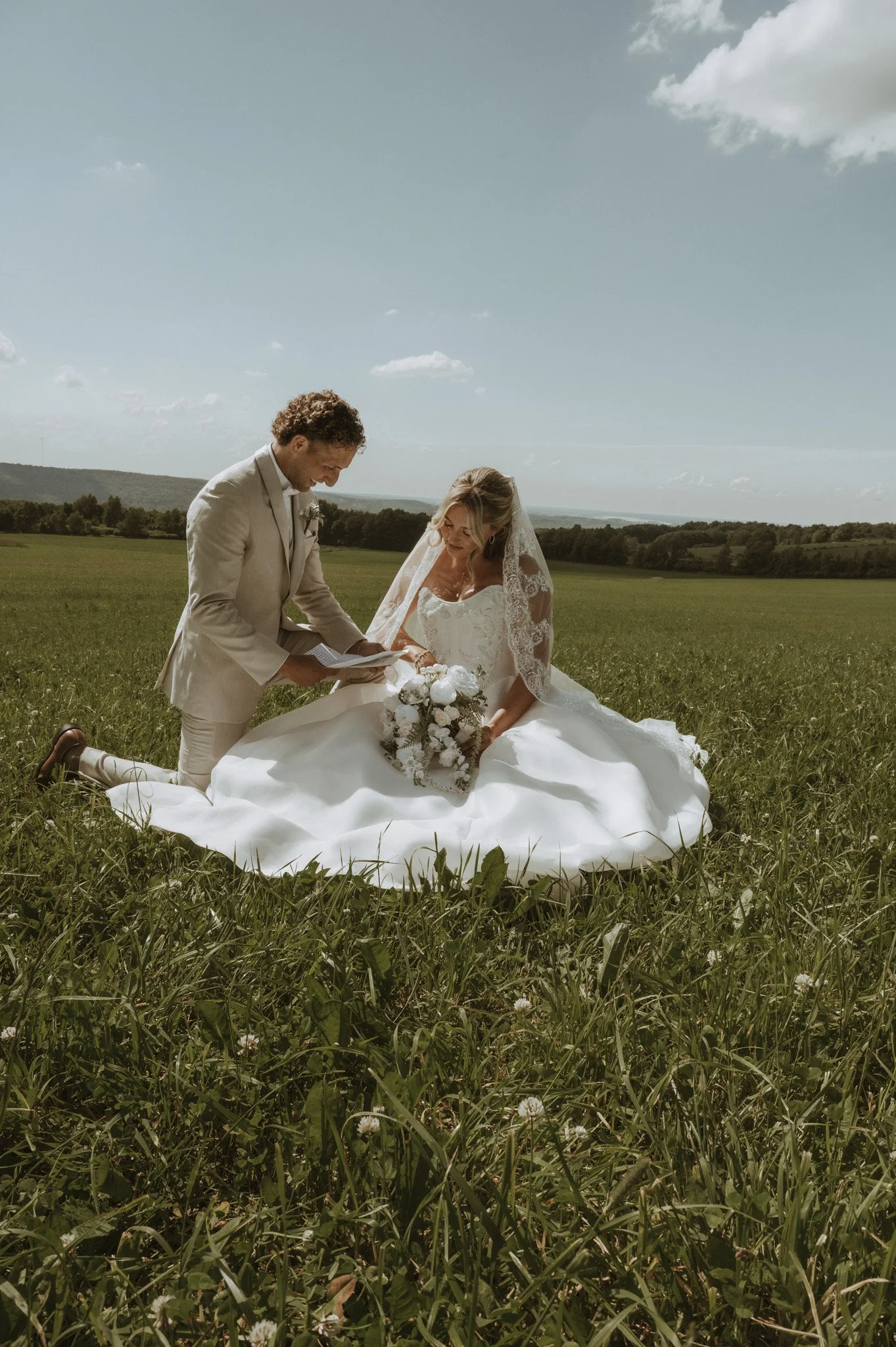 A bride and groom kneeling on the grass in a field, with the groom reading from a paper and the bride holding a bouquet, under a partly cloudy sky.
