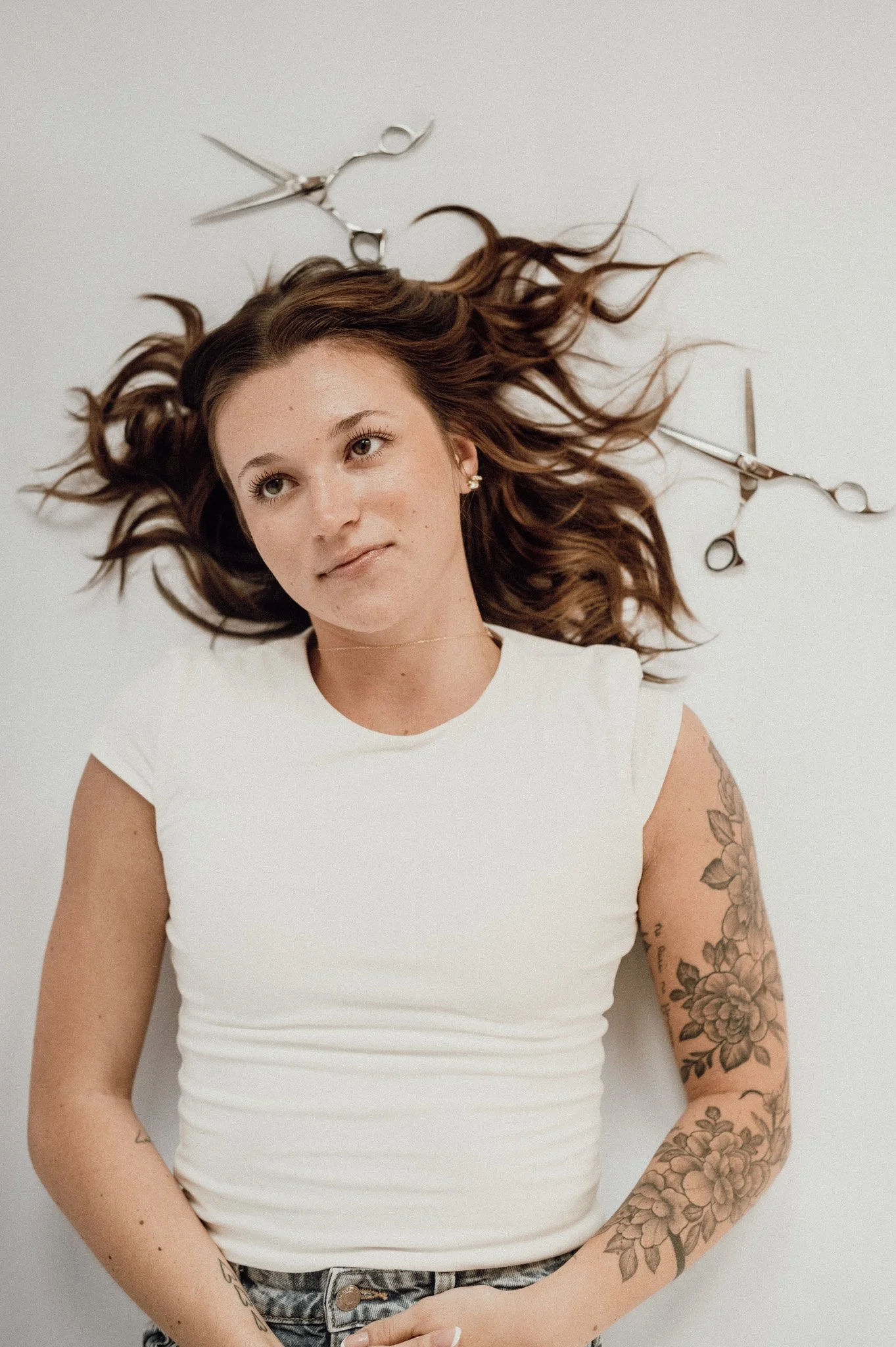 A woman with long brown hair lying on her back on a white surface, holding scissors, wearing a white top and jeans, with tattoos on her arm.