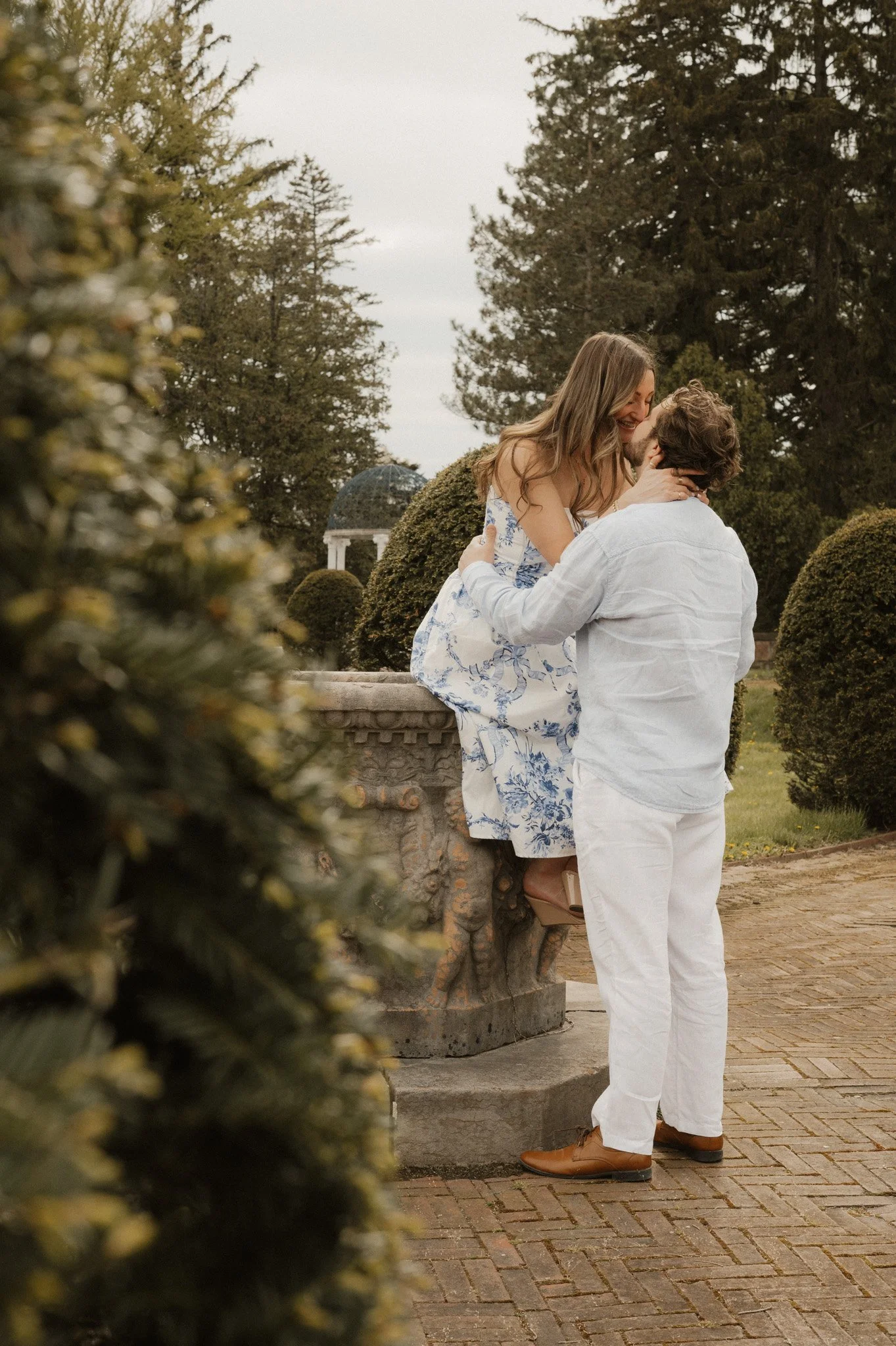 A couple is sharing a kiss outdoors near a stone balustrade, surrounded by neatly trimmed bushes and tall trees, with a pavilion visible in the background.