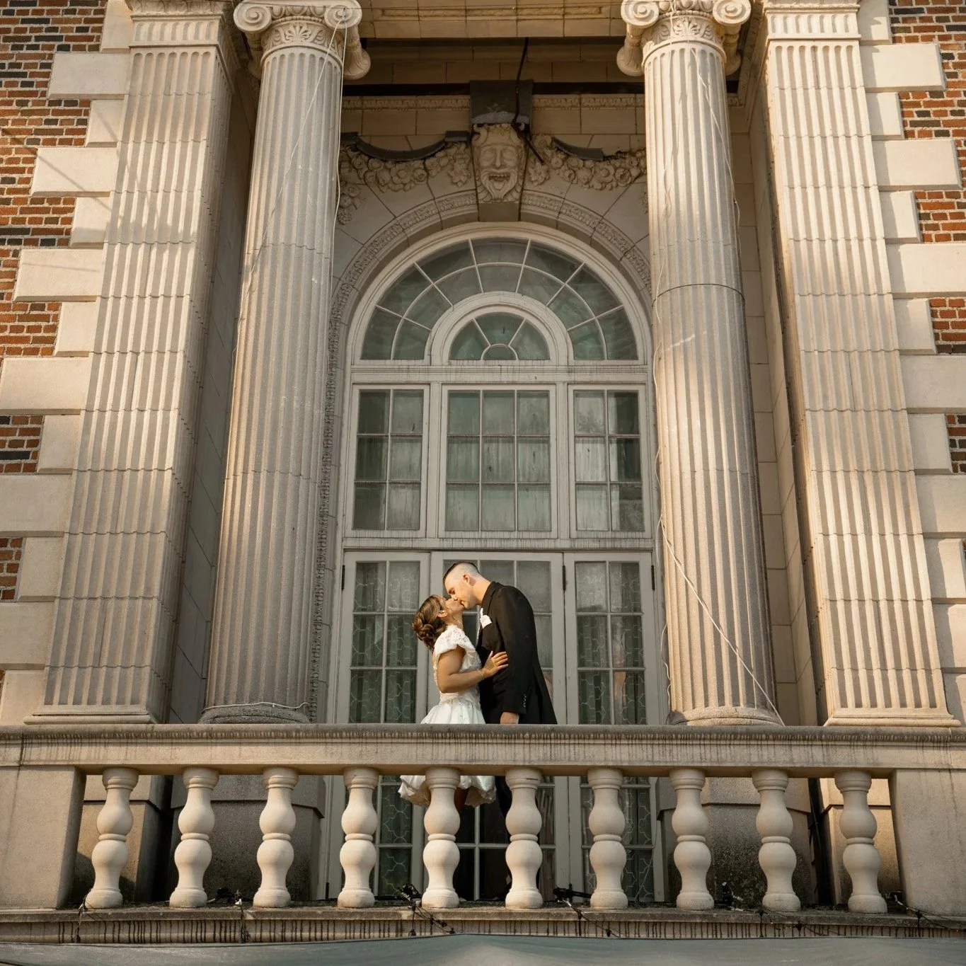 A couple in wedding attire stands on a building's balcony, sharing an intimate kiss under a large arched window with decorative columns and a carved face above.