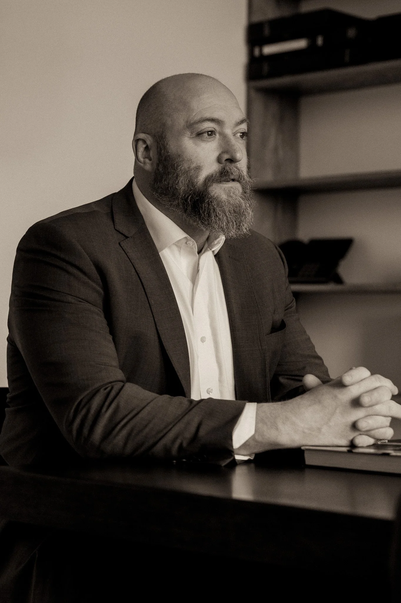 A man with a beard and bald head wearing a suit jacket and white shirt sitting at a desk with folded hands, in a room with shelves in the background.