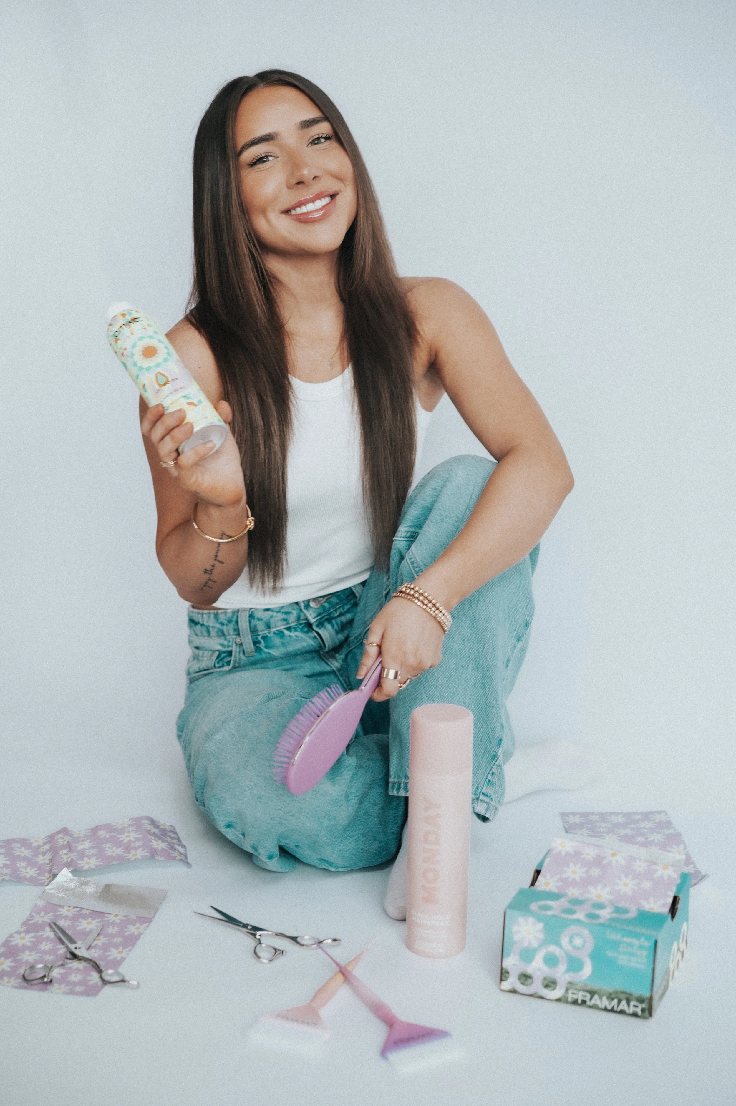 A woman with long brown hair smiling while sitting on the floor surrounded by craft supplies, including scissors, paper, and tissue paper, holding a tube of foam spray and an origami paper animal.