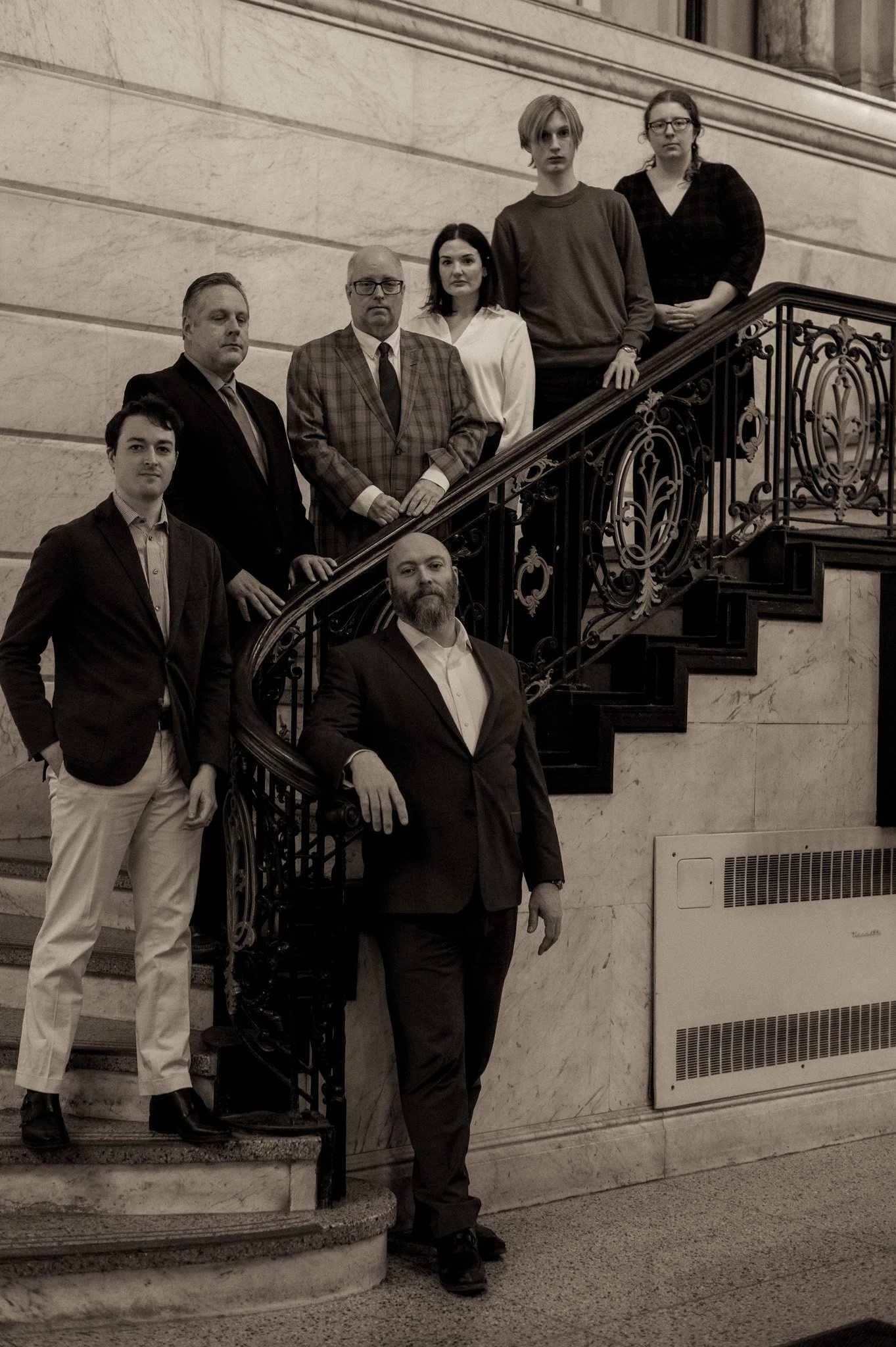 A group of six people standing on a staircase inside a building with marble walls and ornate iron railings. The individuals are dressed in formal and semi-formal attire, and appear to be posing for a photo.