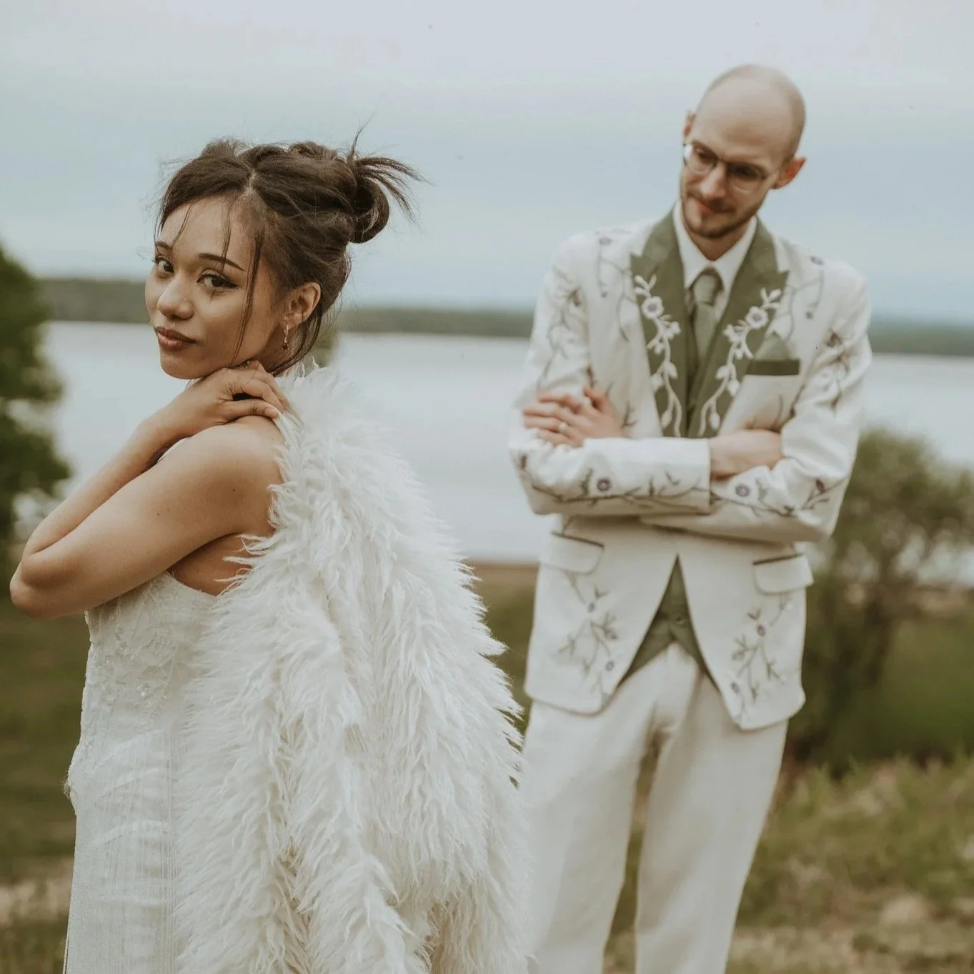 A woman with styled hair and wearing a white dress with a fluffy vest stands outdoors near a body of water, with a man in a white suit with floral embroidery in the background, both posing during daytime.