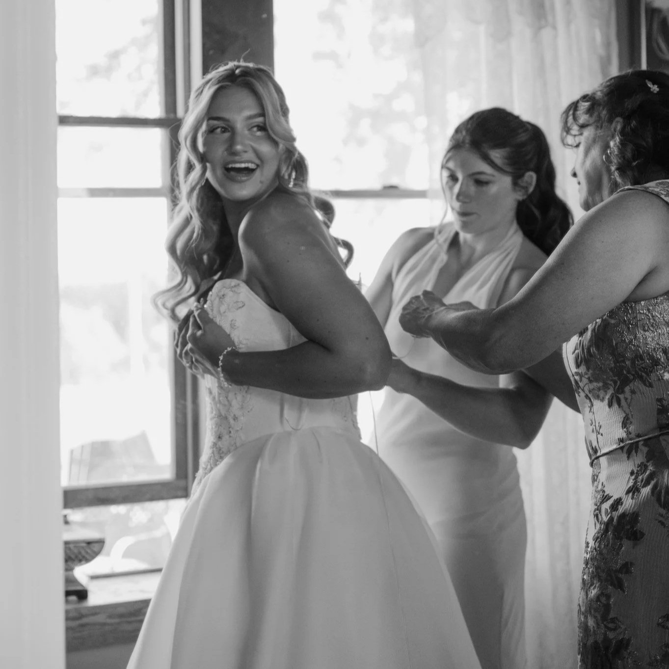 A bride with long wavy hair wearing a strapless wedding dress, smiling as she is helped into her gown by two women, one in a sleeveless dress, inside a room with a window in the background.