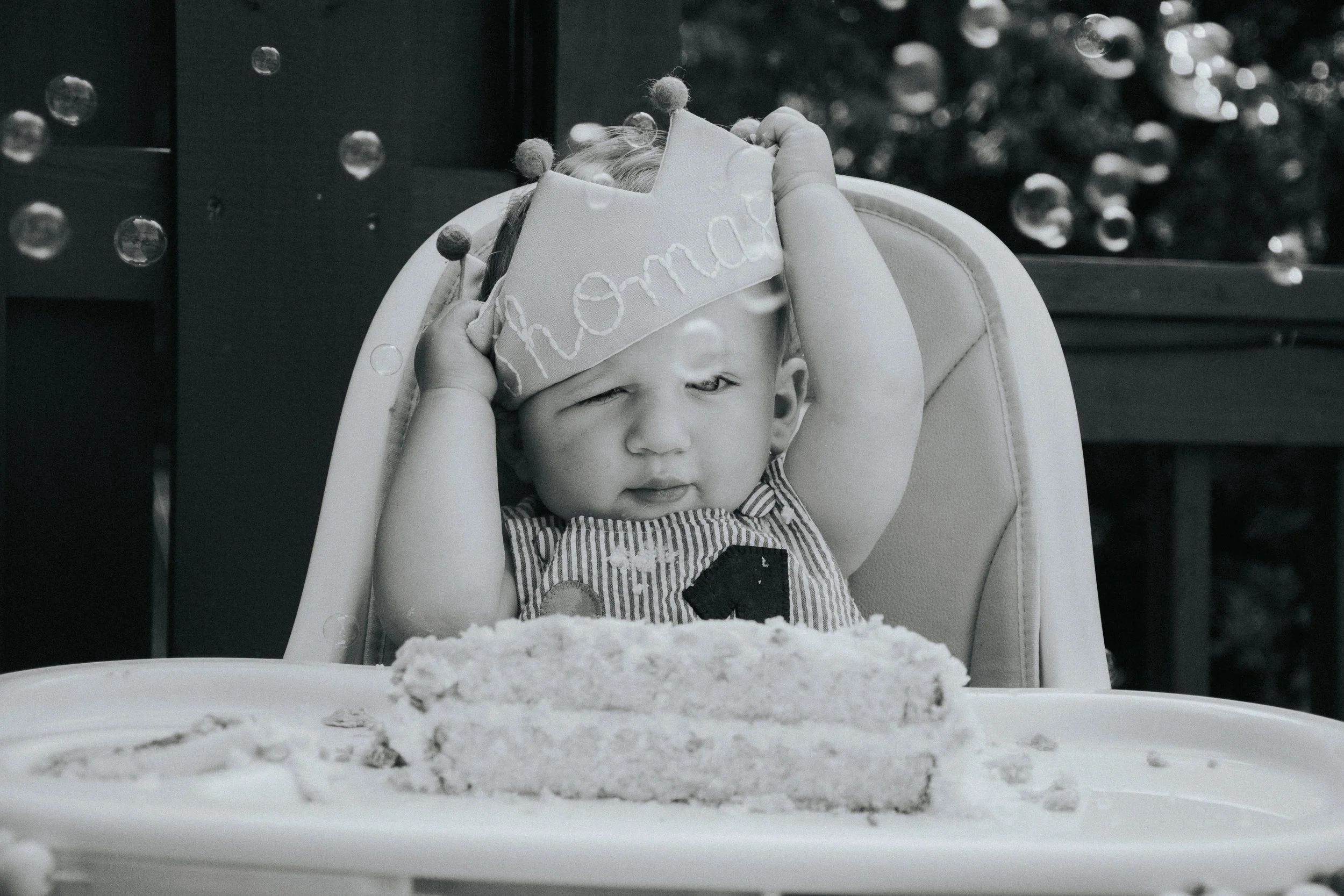 A young child sitting in a high chair with a birthday cake in front, wearing a birthday hat, one eye closed, and holding the hat with hands.