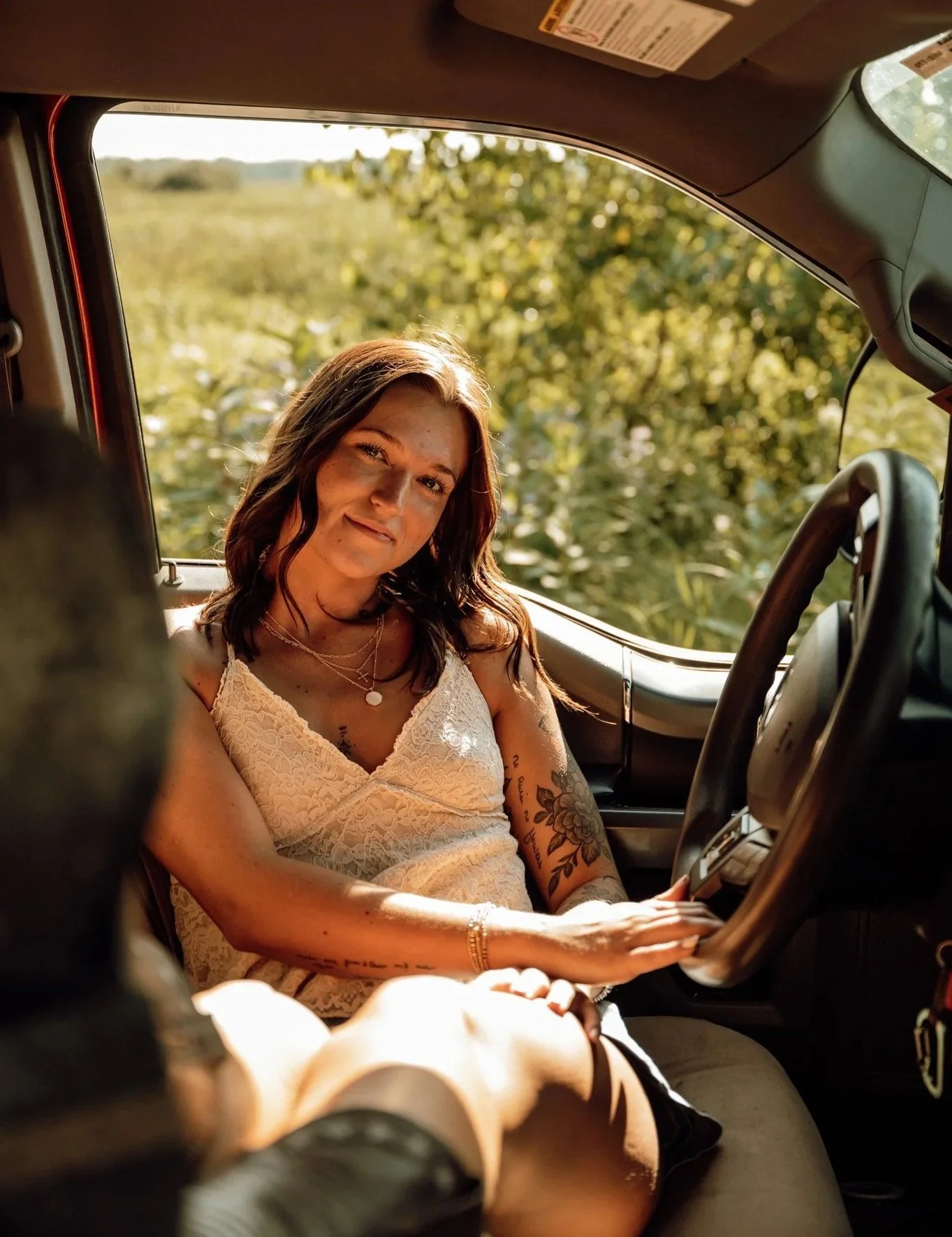 A young woman with wavy brown hair wearing a white lace top sitting inside a vehicle, smiling at the camera, with trees and a fence visible outside the window.