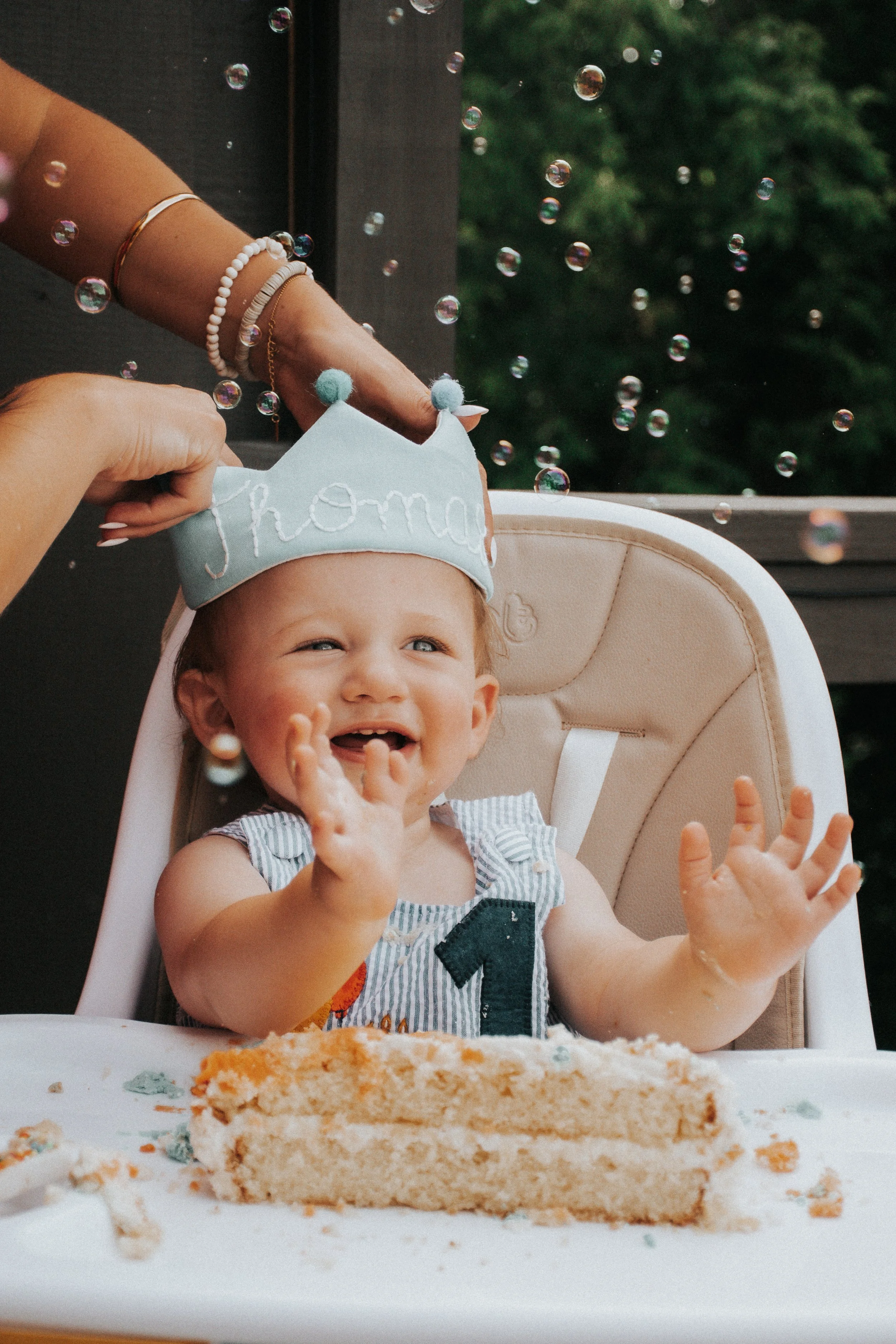 A young child seated in a high chair with a birthday cake in front, wearing a party crown that says 'Mom' and a striped outfit with a number 2 on it. The child is smiling and reaching out with their hands. In the background, there is a blurred outdoor scene with greenery.
