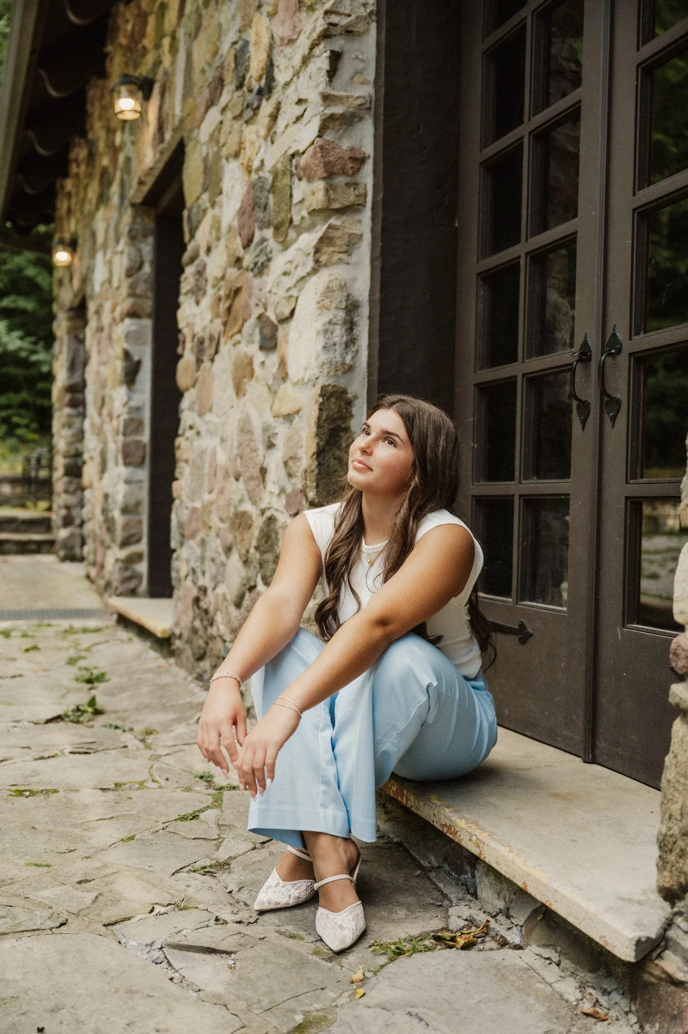 A young woman sitting on the ground outside a stone building with large, dark wooden double doors and black handles. She has long, dark hair and is wearing a white sleeveless top, light blue pants, and white lace shoes. The scene has a relaxed, contemplative atmosphere.