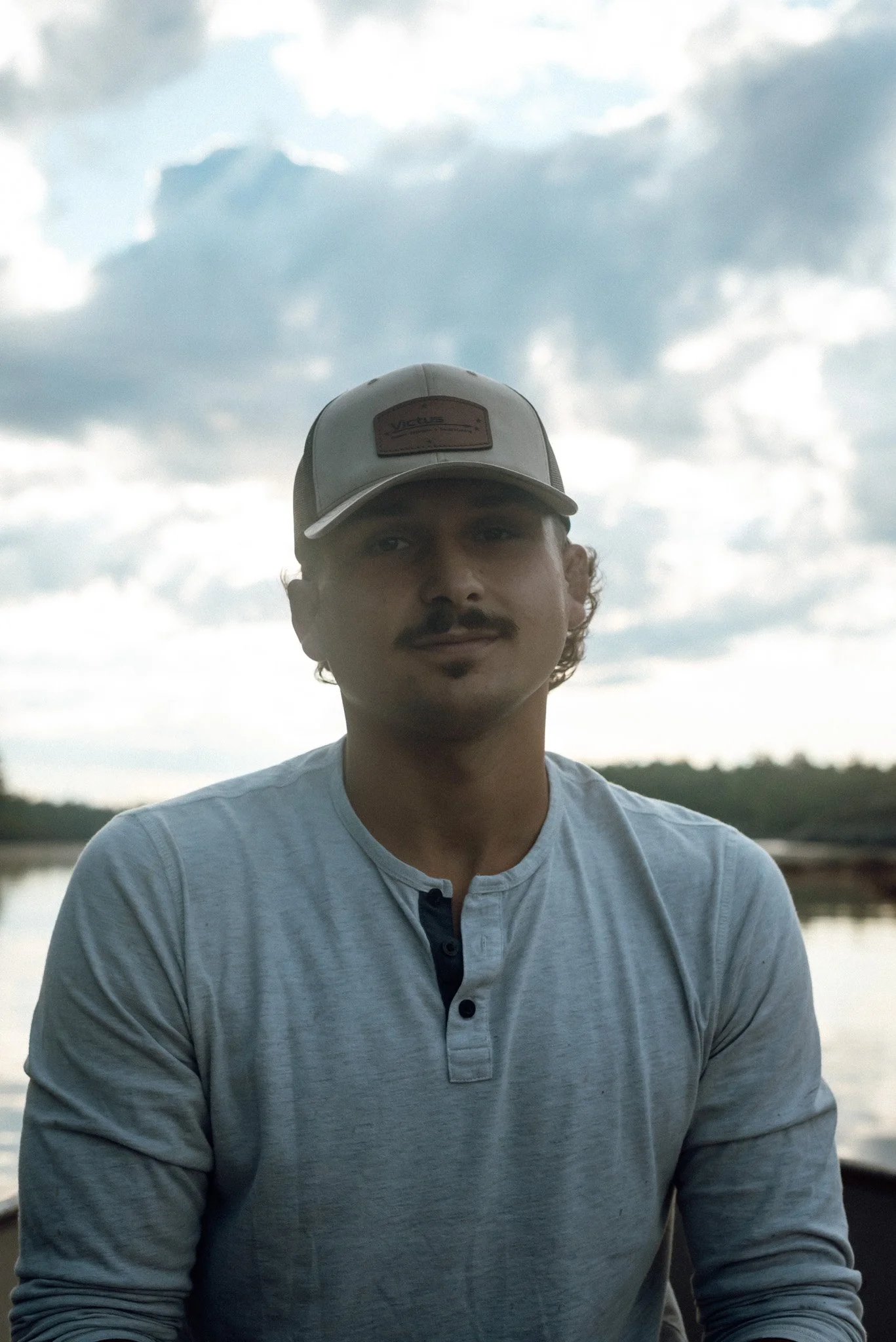 A young man with a light mustache and beard wearing a light-colored baseball cap and a light-colored long-sleeve shirt standing outdoors during daytime, with cloudy sky and trees in the background.