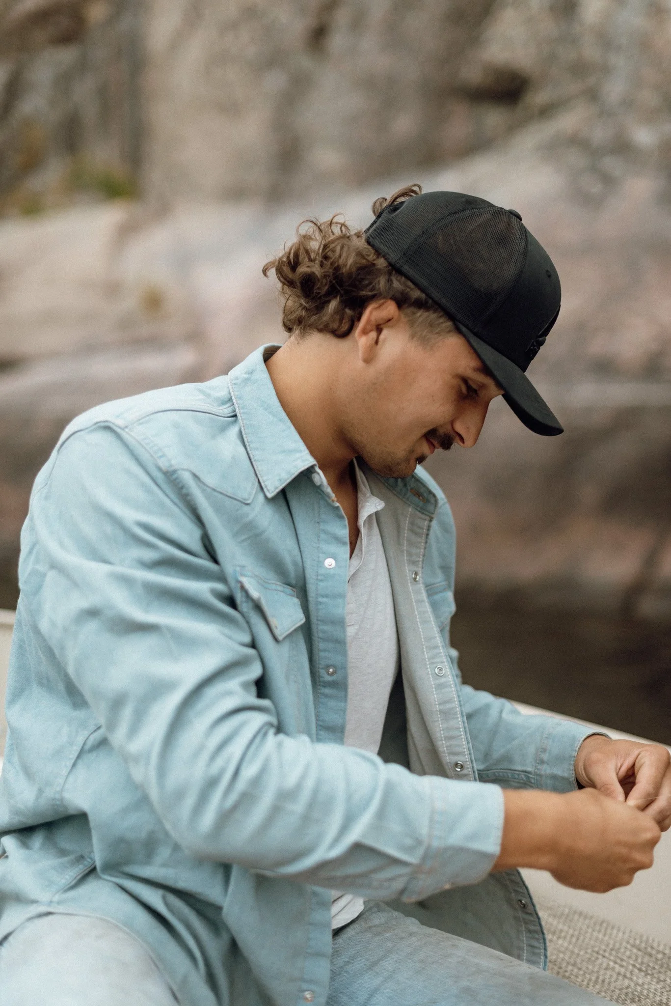 A young man with curly hair wearing a black baseball cap, light blue denim jacket, and white shirt, sitting and looking down, possibly using his phone with a rocky or desert-like background.