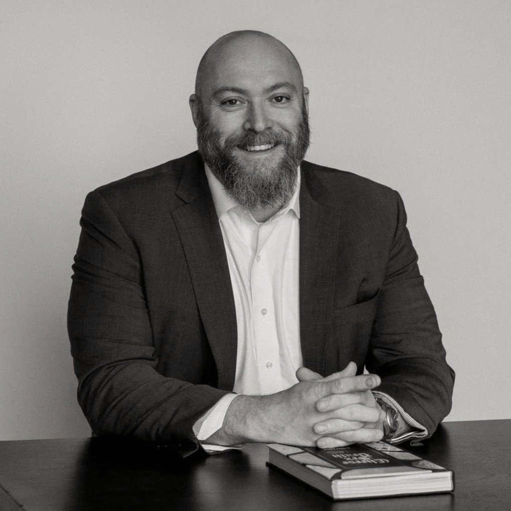 A man with a beard and a shaved head wearing a dark blazer and a white shirt, sitting at a table with his hands clasped, smiling at the camera, with a closed book on the table.