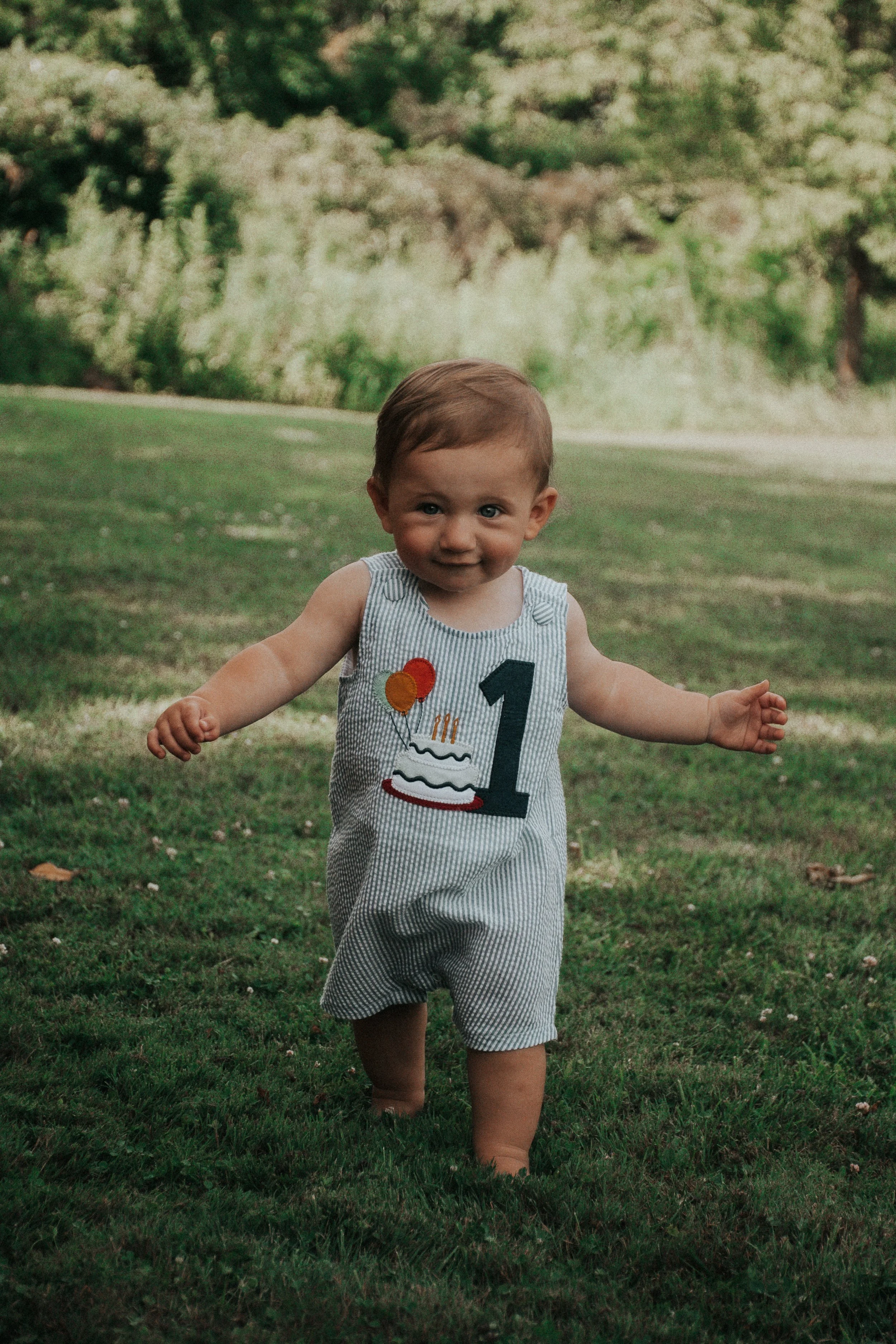 A young child with short brown hair and blue eyes is standing barefoot on a grassy lawn, smiling, and wearing a sleeveless outfit with the number 1 and a birthday cake with balloons embroidered on it, outdoors with a background of green trees.