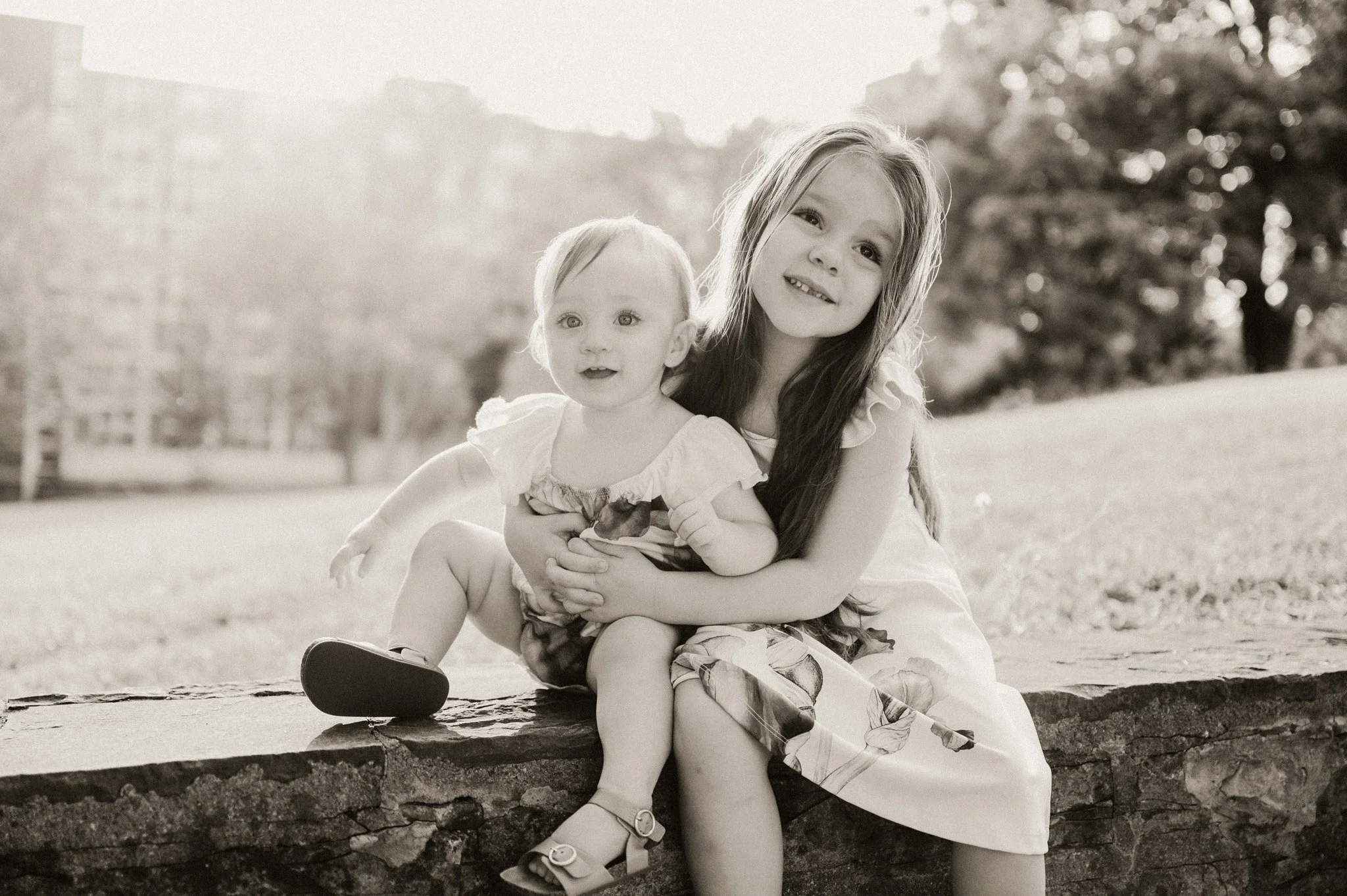 Two young girls sitting on a low stone wall outdoors, smiling and looking at the camera, with trees in the background. It appears to be taken in a park during daylight.