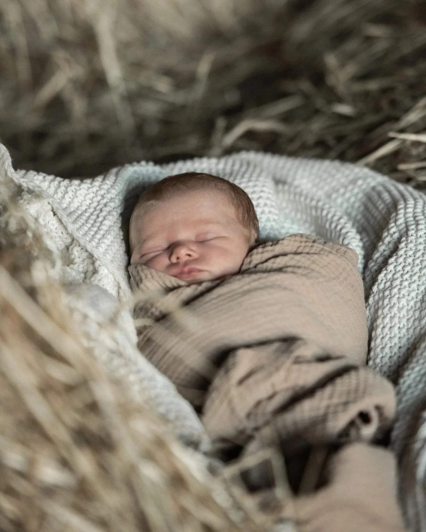 Close-up of a sleeping newborn baby wrapped in a beige and brown striped blanket, lying on a white knit blanket inside a woven basket.