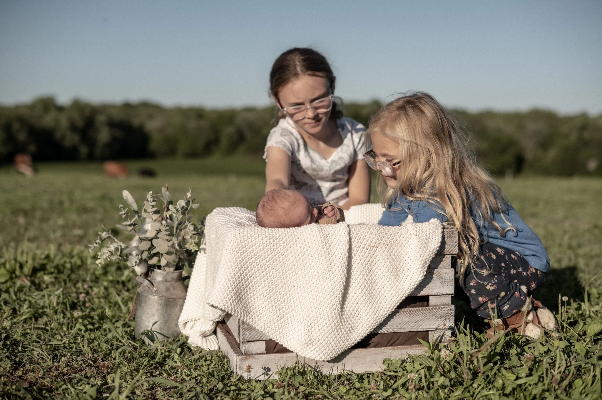 Two young girls with glasses and long hair are outdoors on a grassy field, leaning over a wooden box with a baby doll inside. The girl on the left with dark hair is wearing a grey patterned top, and the girl on the right with blonde hair is wearing a blue top and a floral skirt. The scene includes a bouquet of greenery and flowers placed nearby.