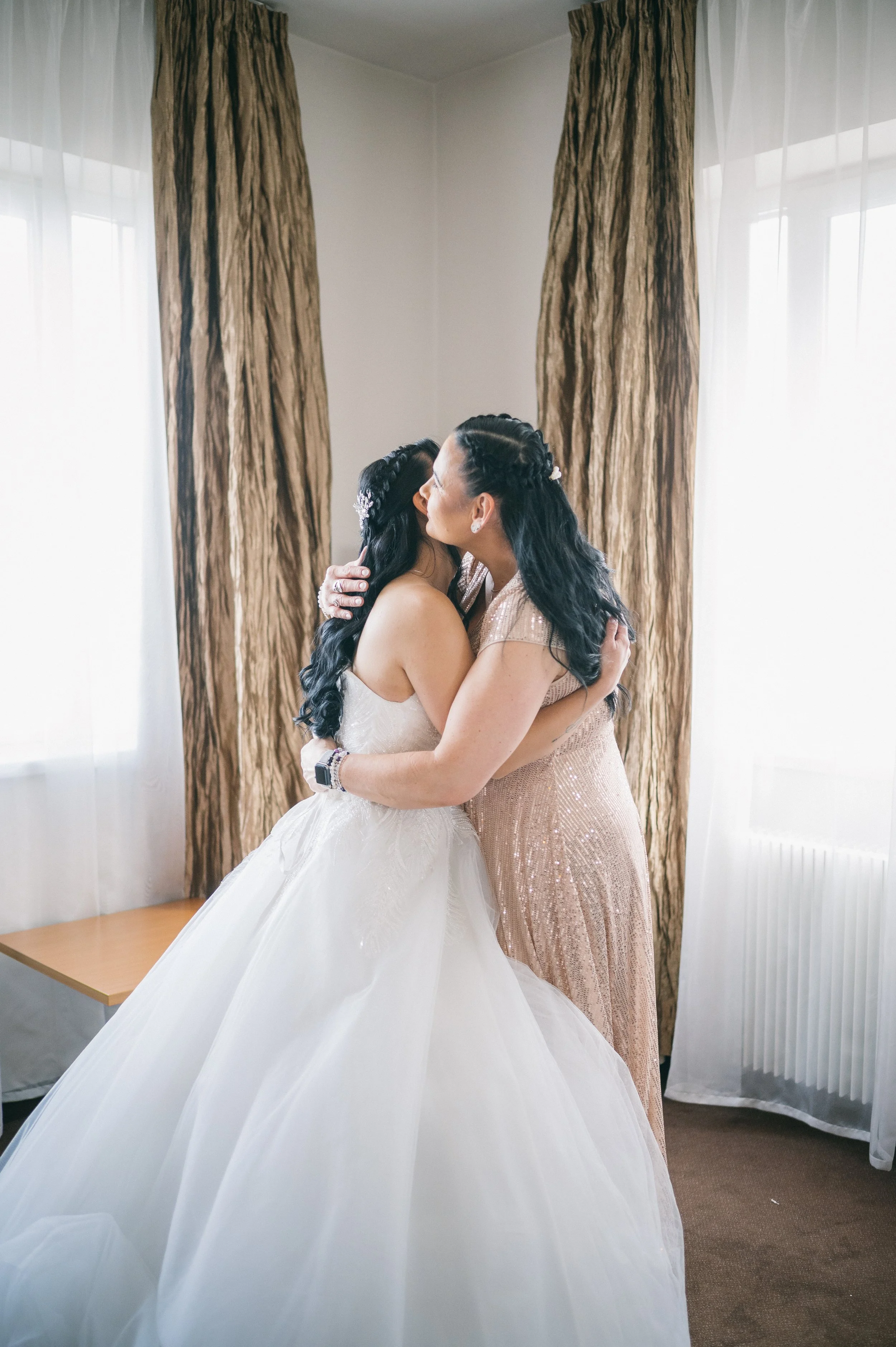 A man with light skin and a shaved head sitting on a gray chair, putting on a brown dress shoe while wearing a white shirt and beige pants. The room has gold-patterned curtains and a lamp in the background.