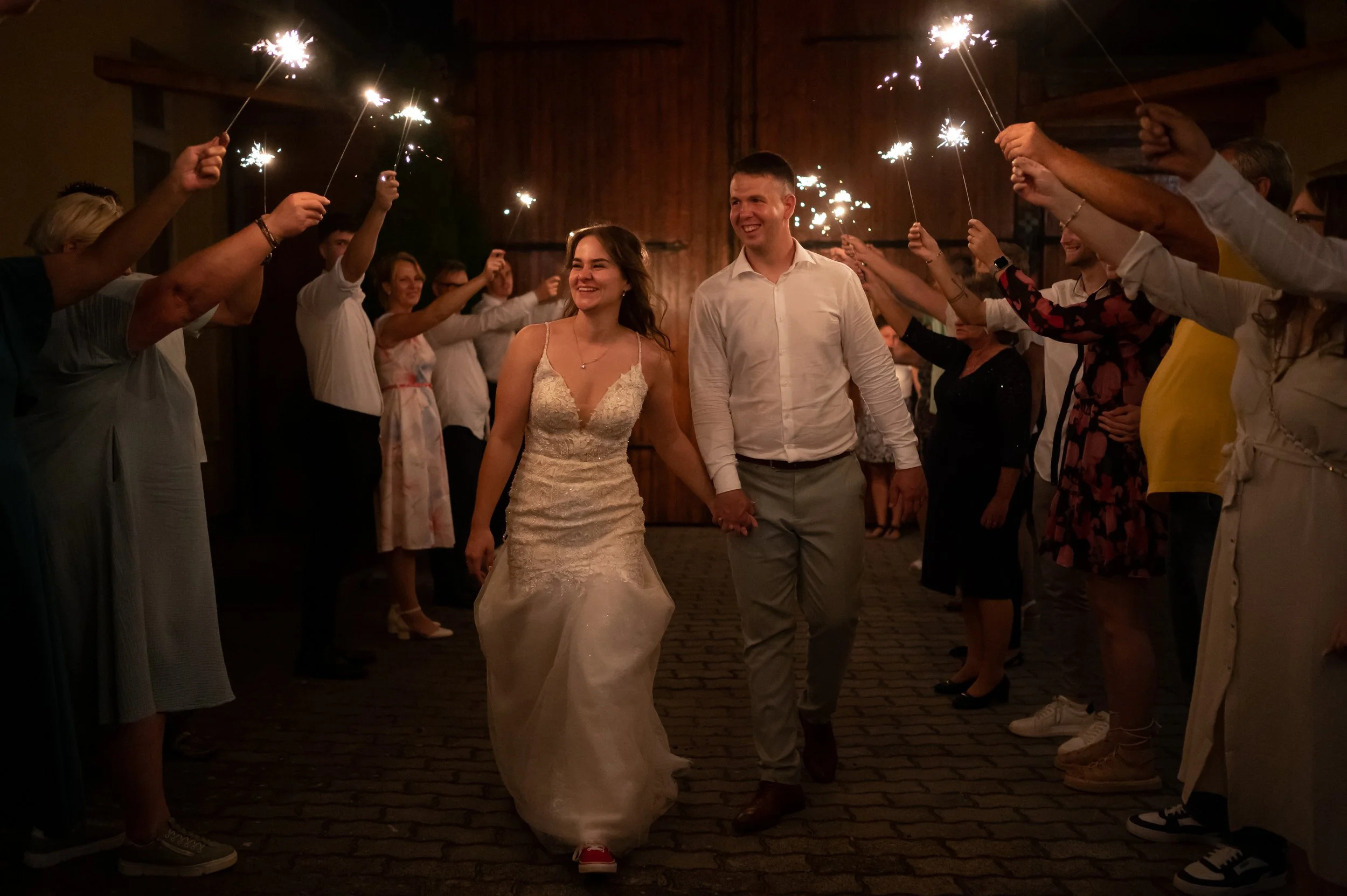 A bride smiling and looking down, with two women beside her assisting her into her wedding dress, in a bright room with curtains.