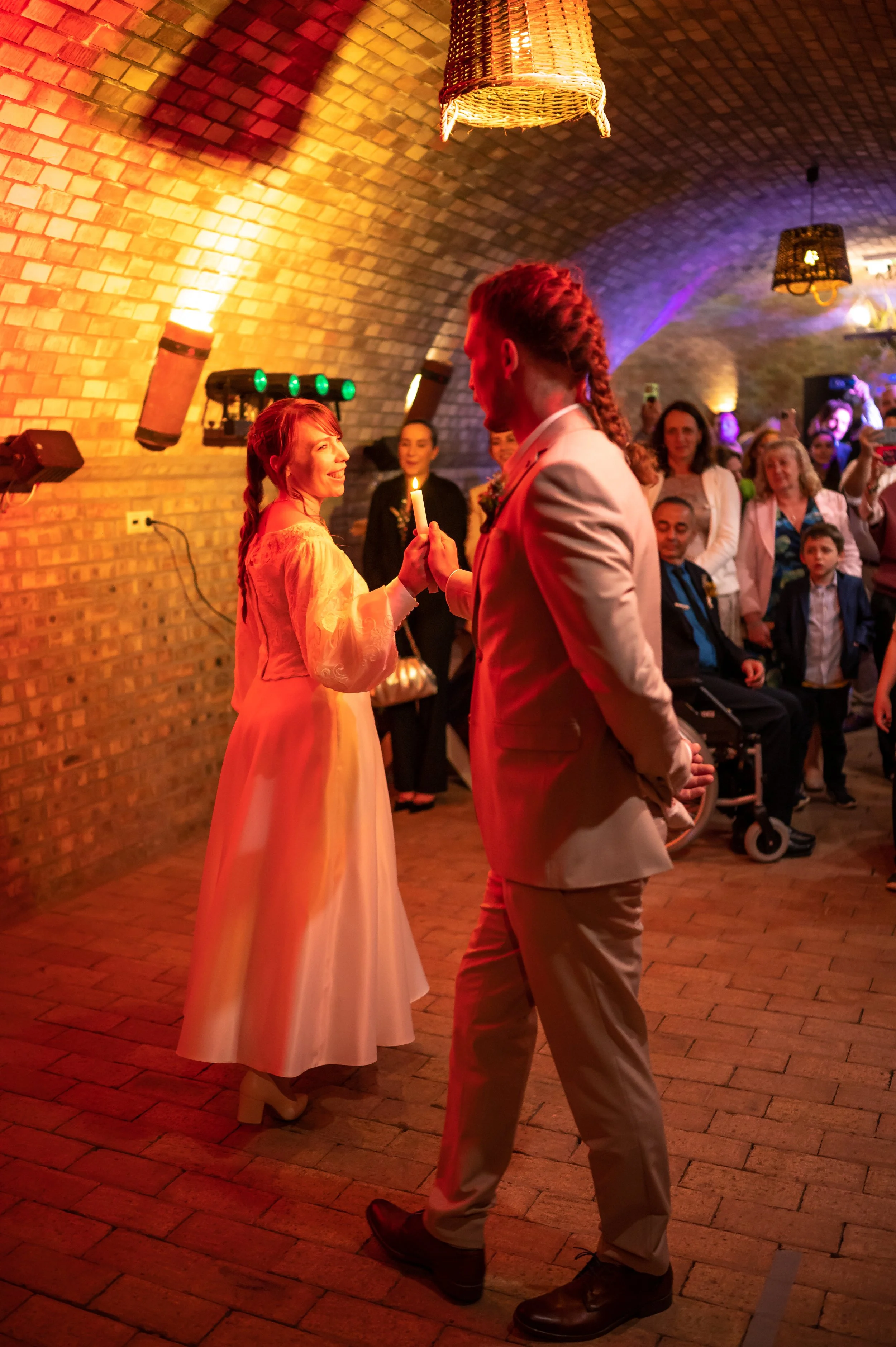 A bride in a wedding gown is being assisted by two women with the gown outside the bed. The bride is holding a veil, and the room has a framed picture, a mirror, and furniture.
