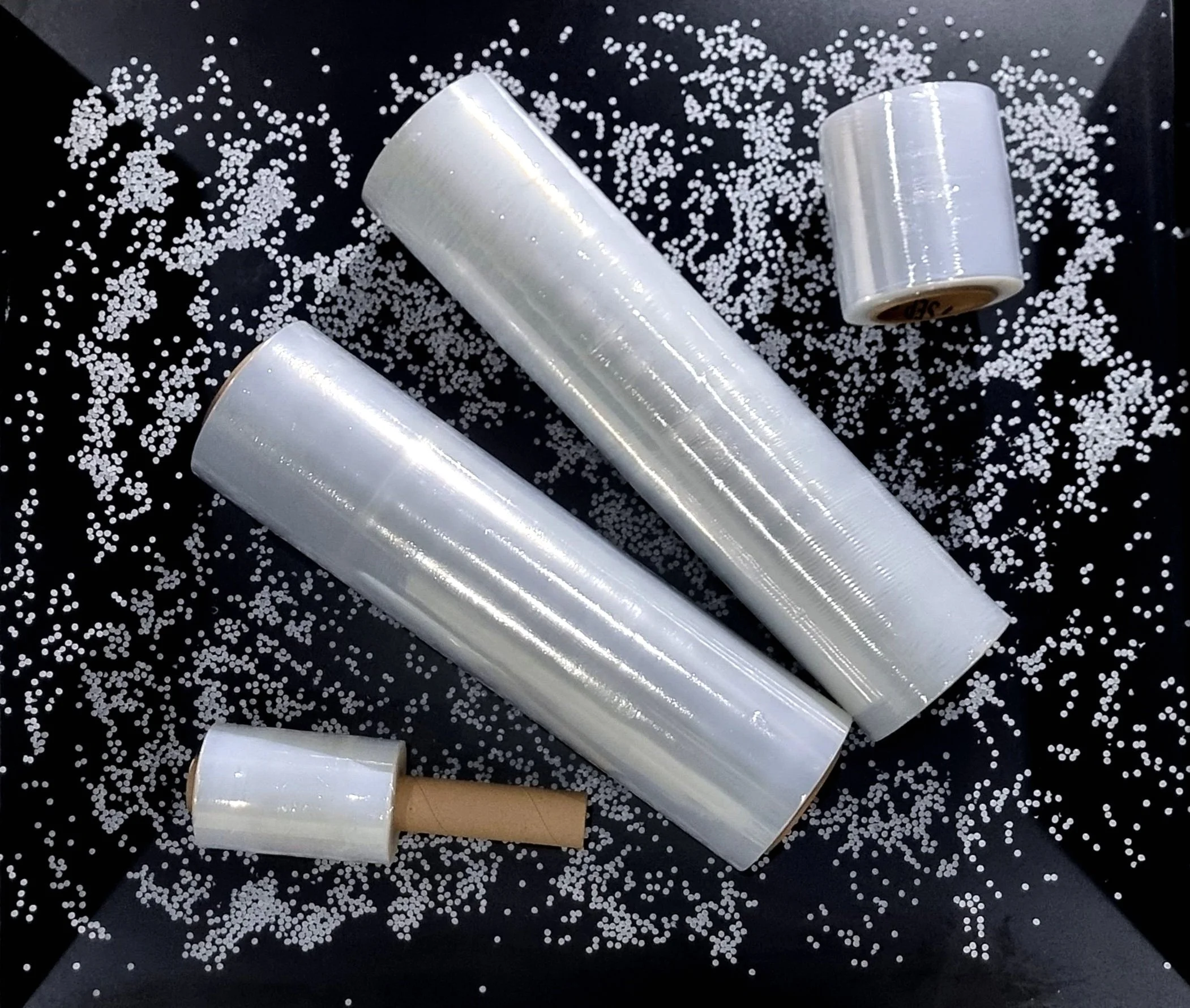 Rollos de plástico transparente, un rodillo y pequeñas bolitas blancas dispersas sobre un fondo negro.