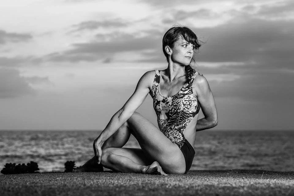 A woman performing a yoga pose on a grassy area near the ocean at sunset.