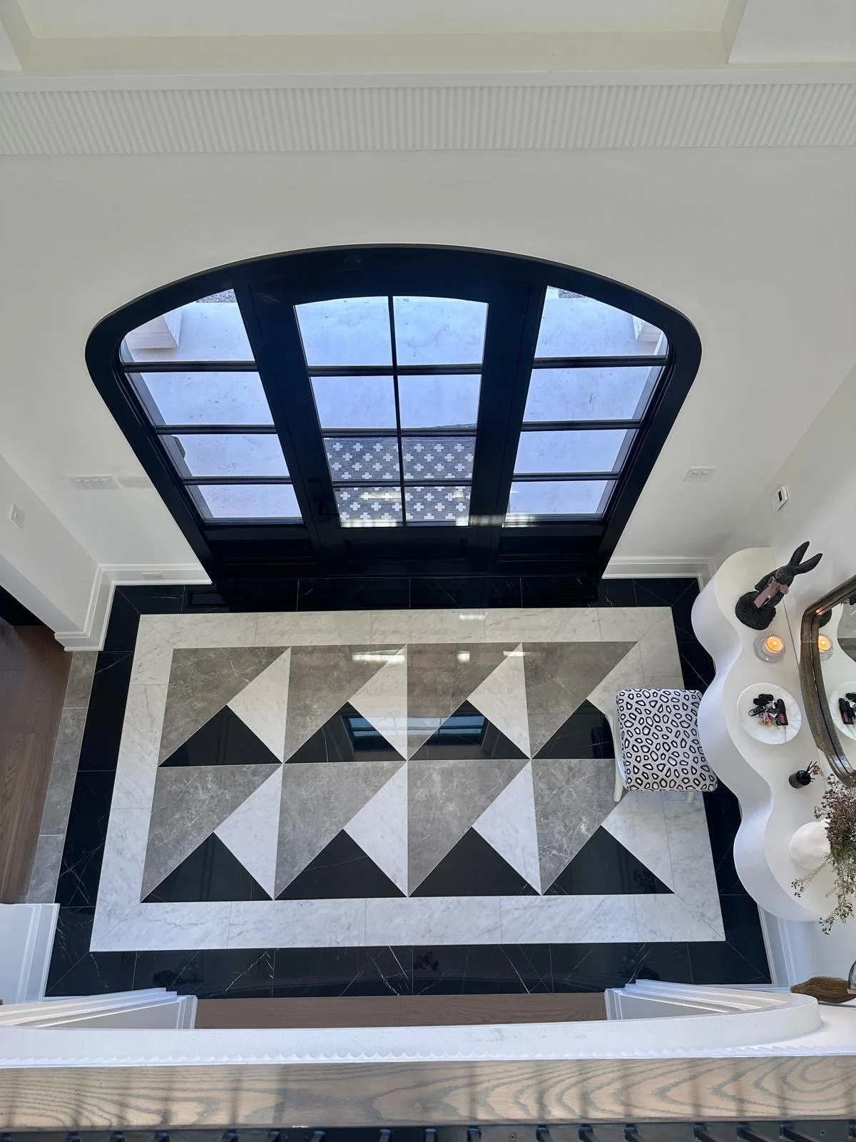 View from above of an entryway with black-framed glass double doors, a patterned tile floor with black, white, and beige geometric shapes, a white wavy console table with decorative items, and a small black and white patterned stool.