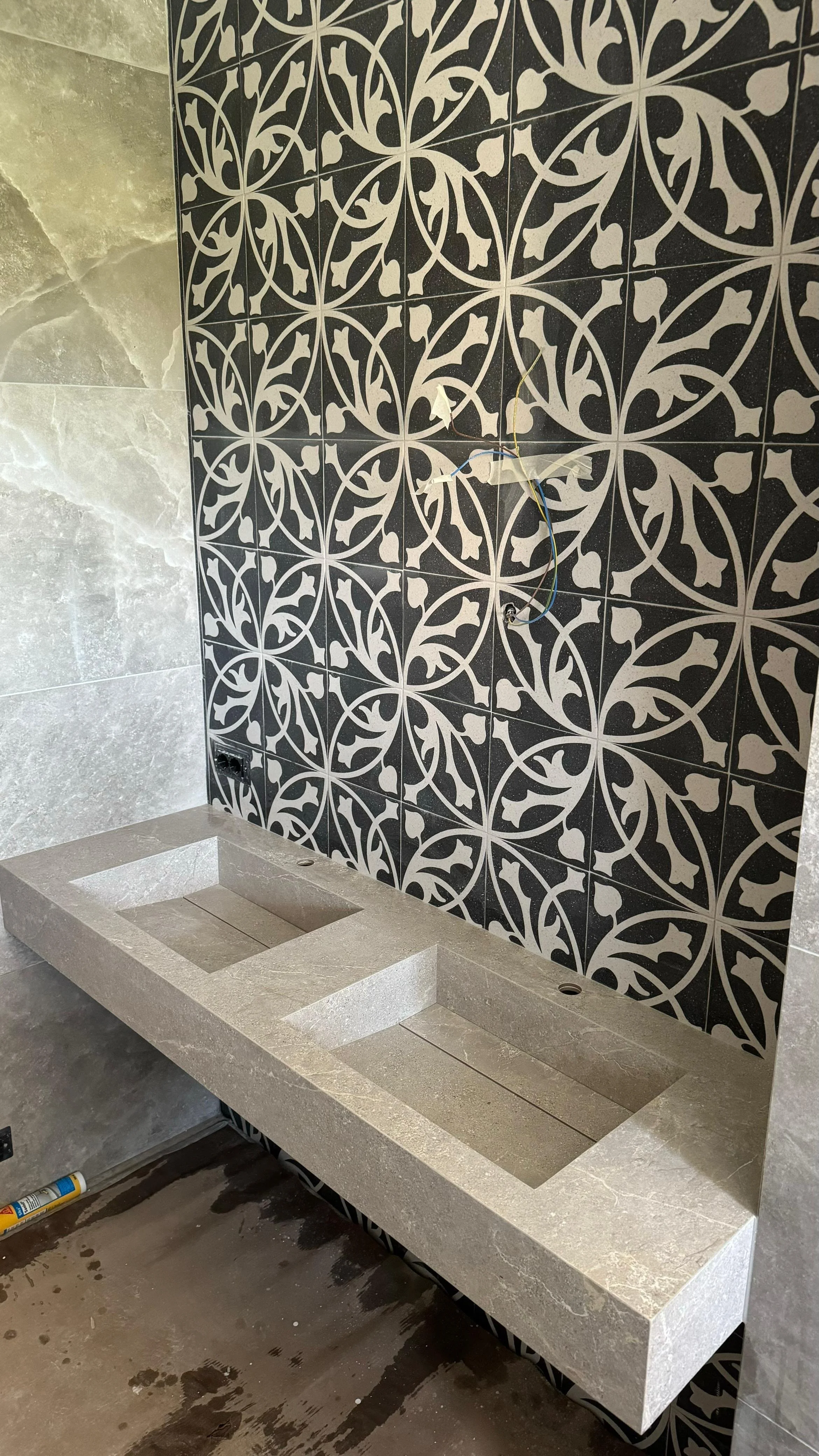 Countertop with two sink cutouts and a decorative black and white patterned tile wall in a bathroom under construction.