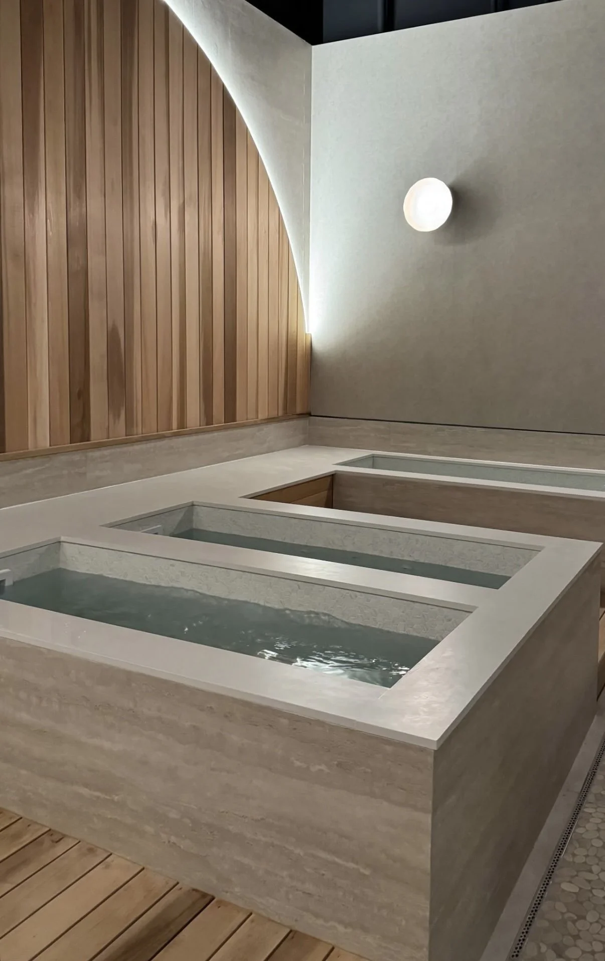 Interior of a modern spa or wellness center with a large stone soaking tub filled with water, wooden flooring, and a mix of wooden and grey walls, illuminated by a natural light feature and a wall-mounted light.