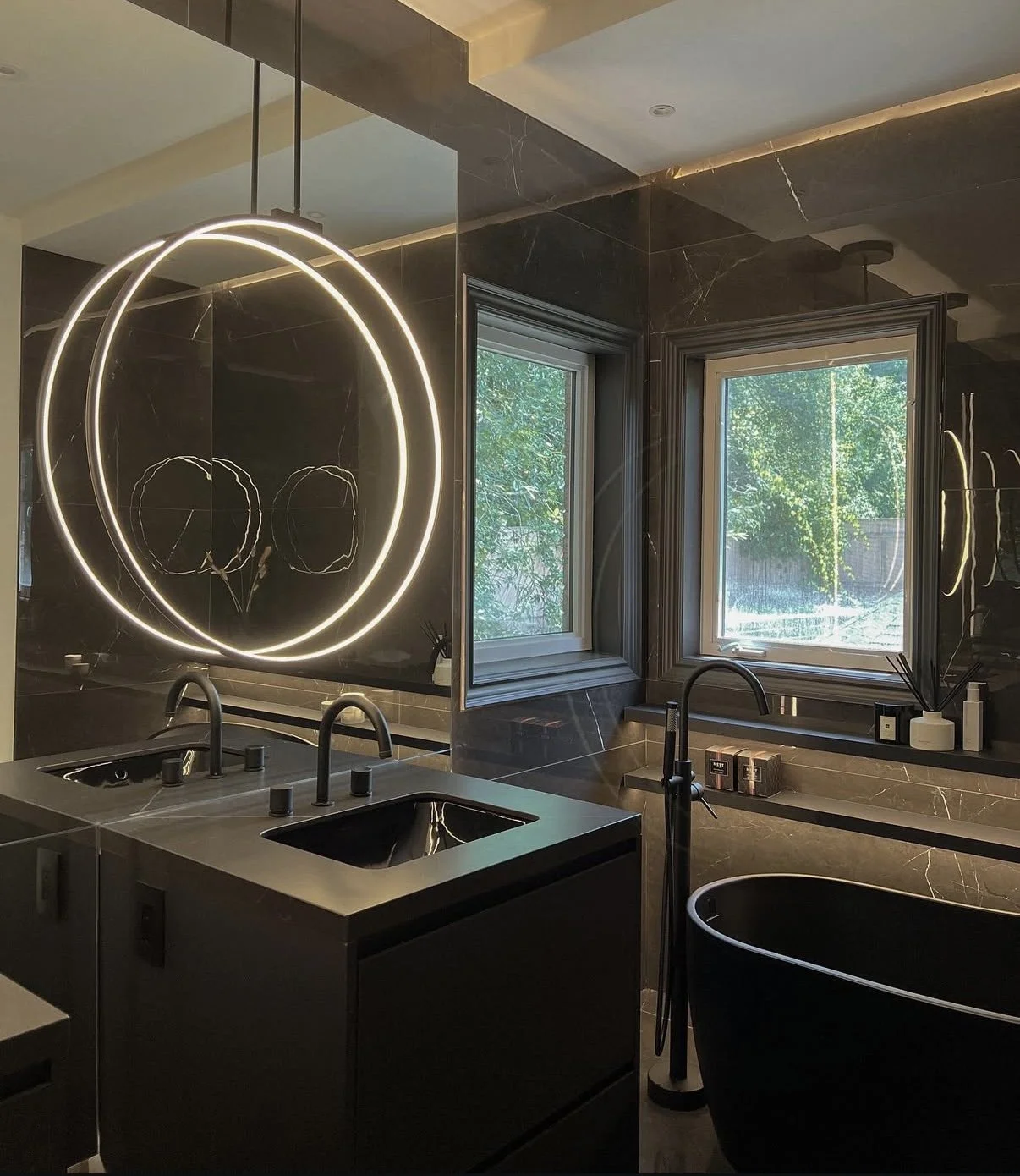 Modern bathroom with black French bathtub, double sinks, black marble walls, circular LED mirror, two windows with green trees outside, toiletries and decorative items on the window sill.