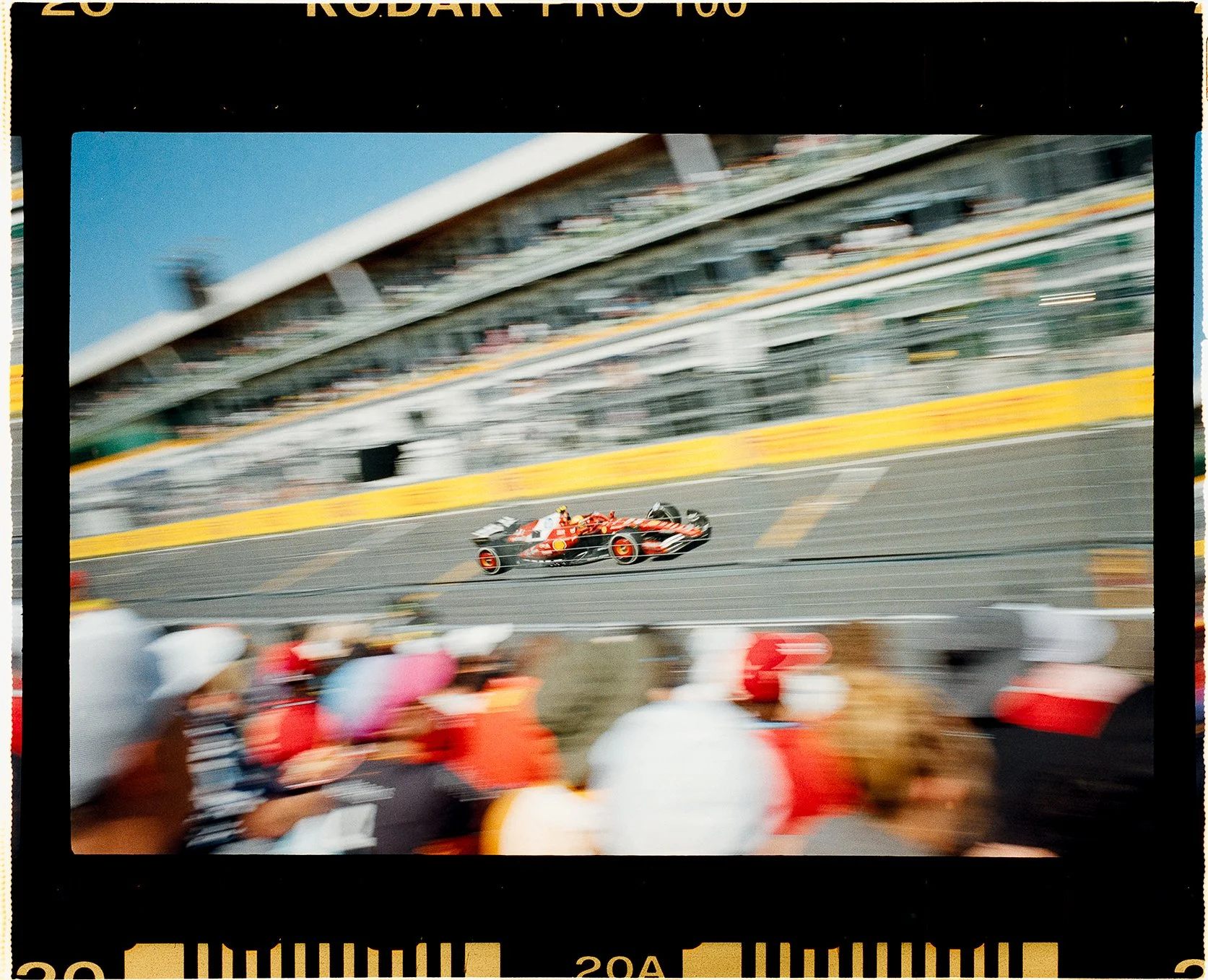Fine art print of Lewis Hamilton's Ferrari at a Formula 1 Grand Prix on the  racetrack with spectators in the foreground and stands in the background, captured in motion blur on 35mm film leica m6