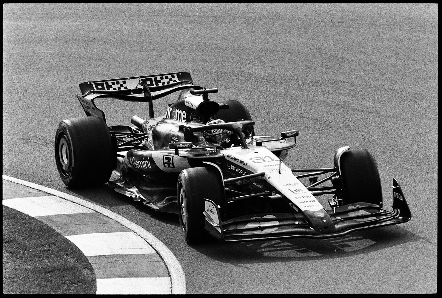 Black and white photograph of a Oscar Piatri's Mclaren racing on a track, taking a turn near the edge marked with striped curbing on 35mm film