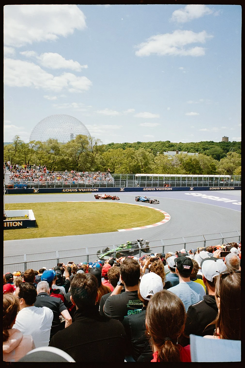 A Formula 1 race in progress with two cars racing on a track surrounded by spectators, trees, and a large geodesic dome in the background.35mm film leica m6