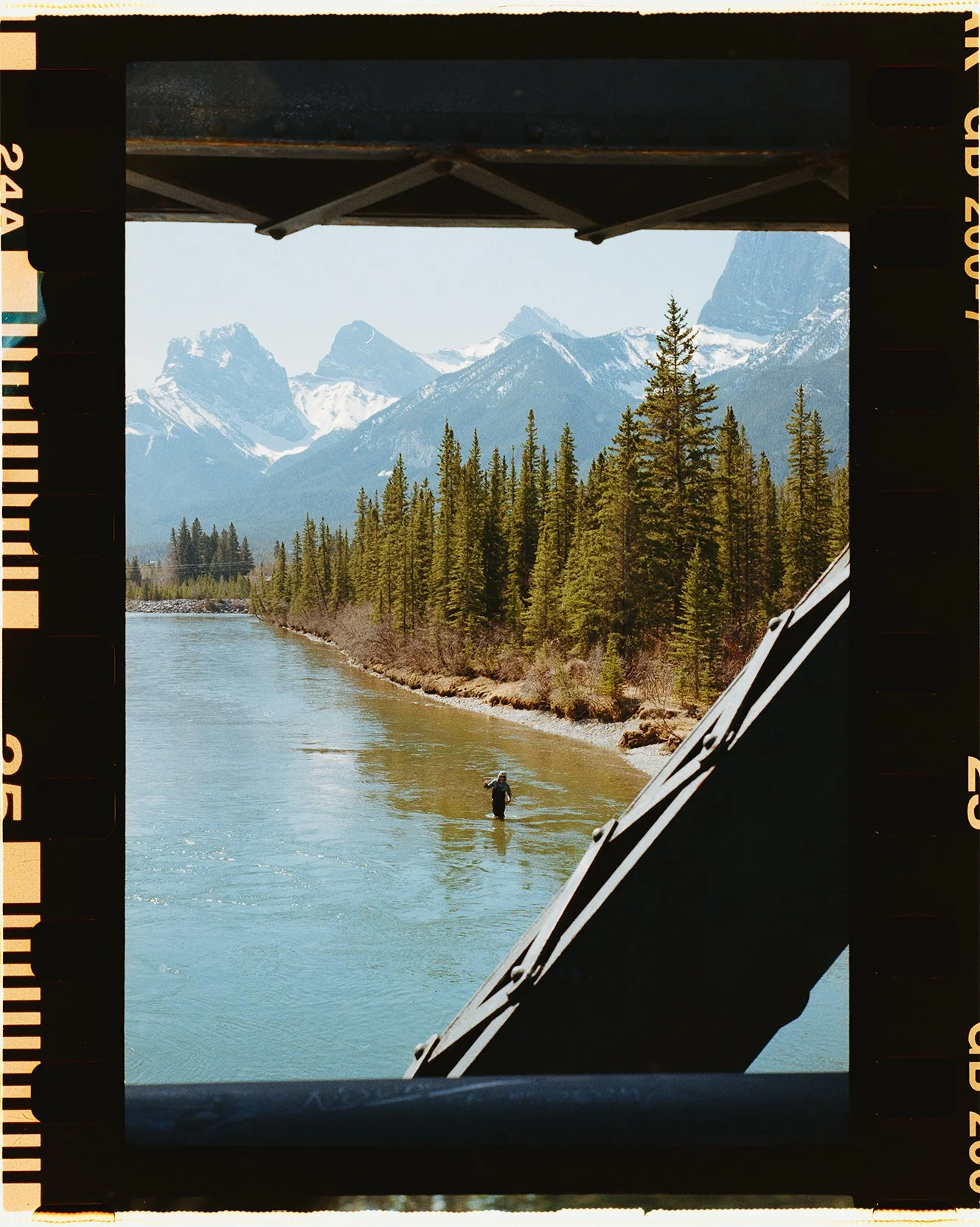 Scenic view of snow-capped Three Sister's in Canmore Alberta, evergreen trees, and a person fishing  in the river, framed by the edge of a bridge on 35mm film leica m6
