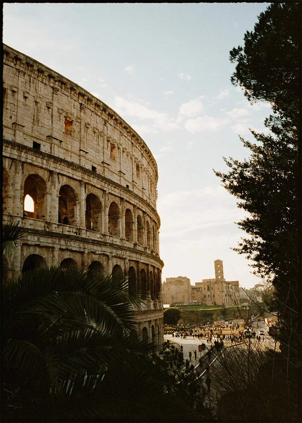 Fine art print of The Colosseum in Rome, viewed during sunset, with trees and buildings in the background and pedestrians on the street on 35mm film Pentax 17